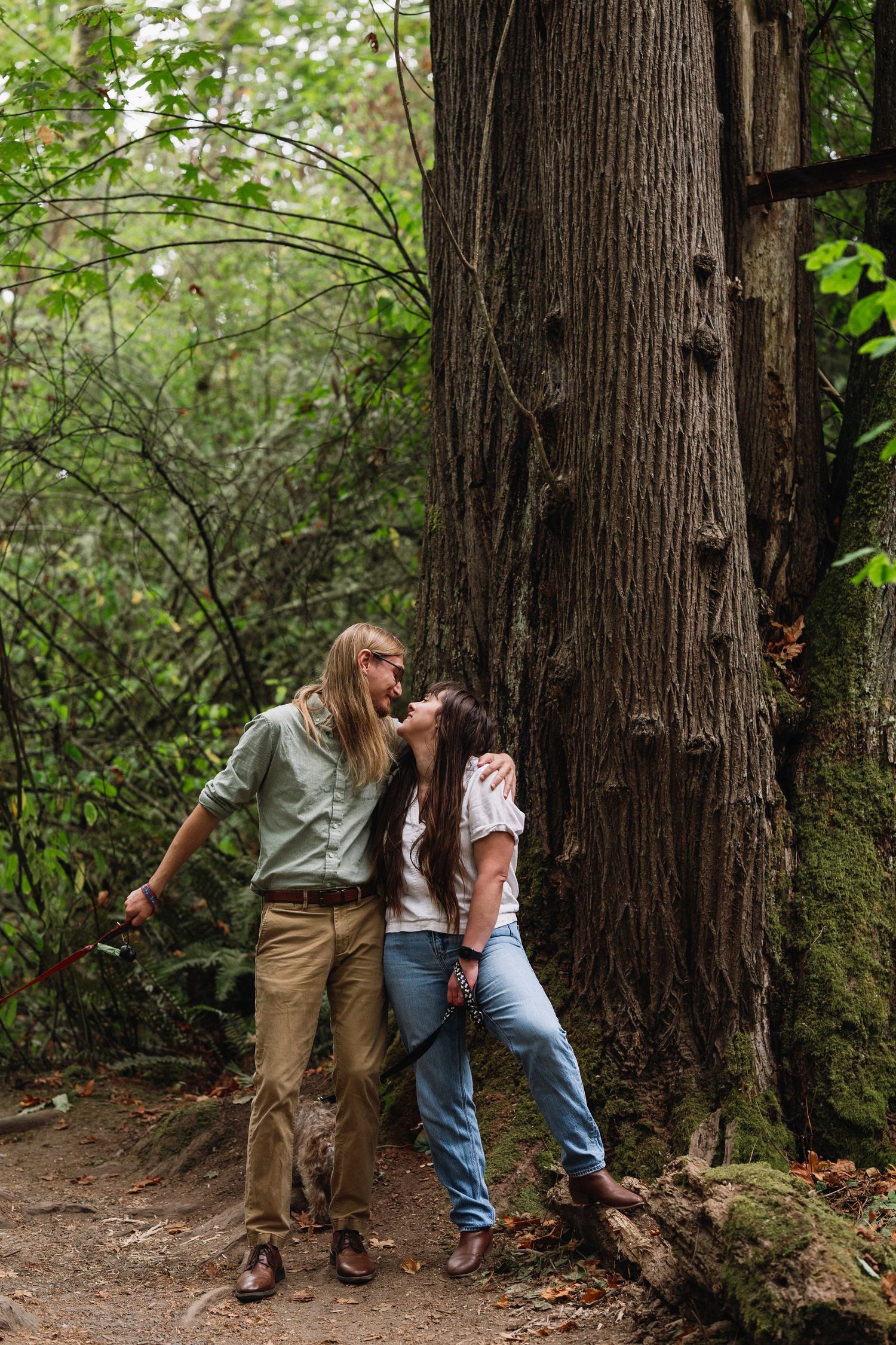 A couple shares a kiss leaning against a massive tree trunk in a lush green forest.