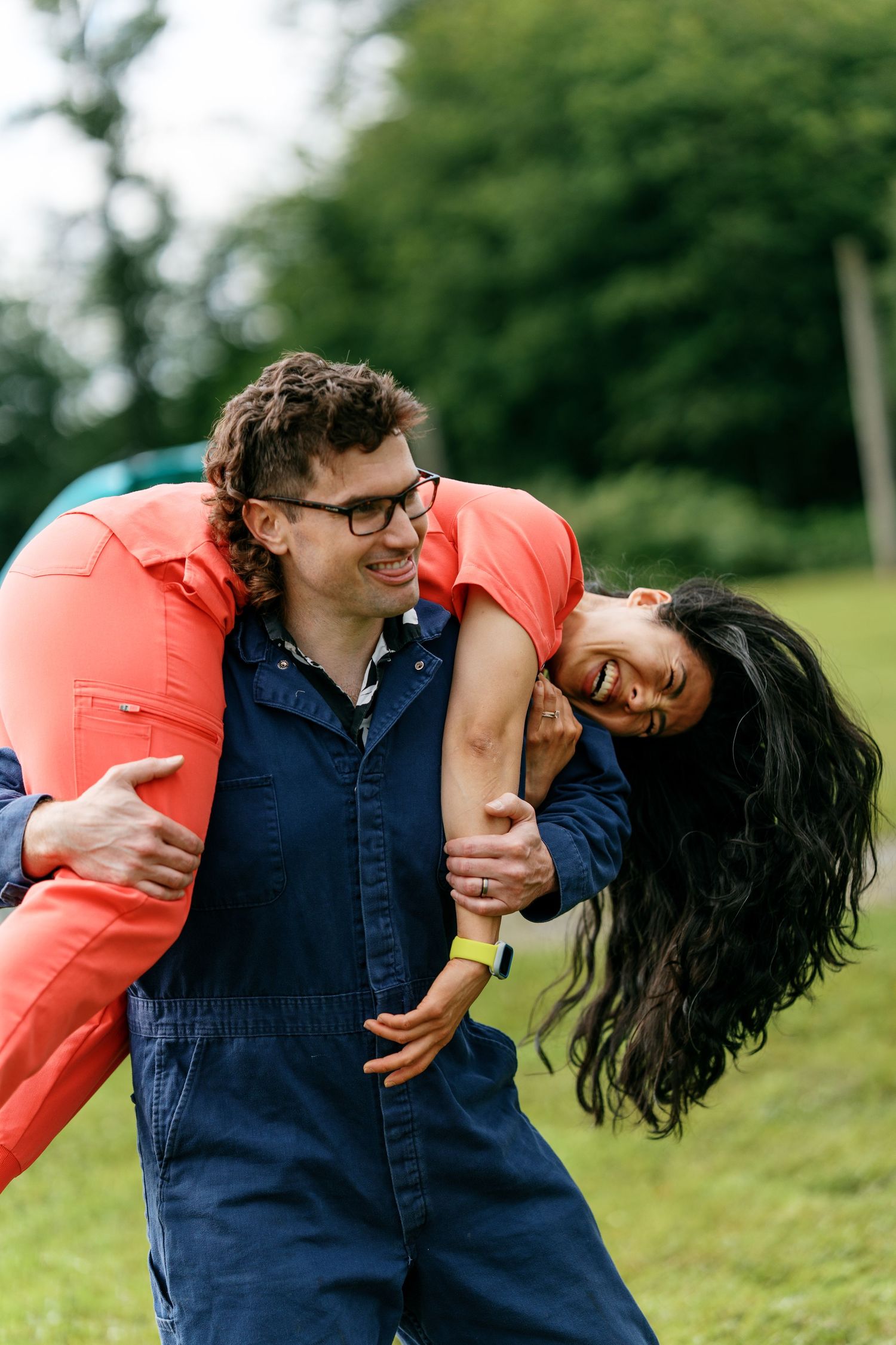 Laughing couple playfully embracing outdoors, man carrying woman over his shoulder in a green park setting.