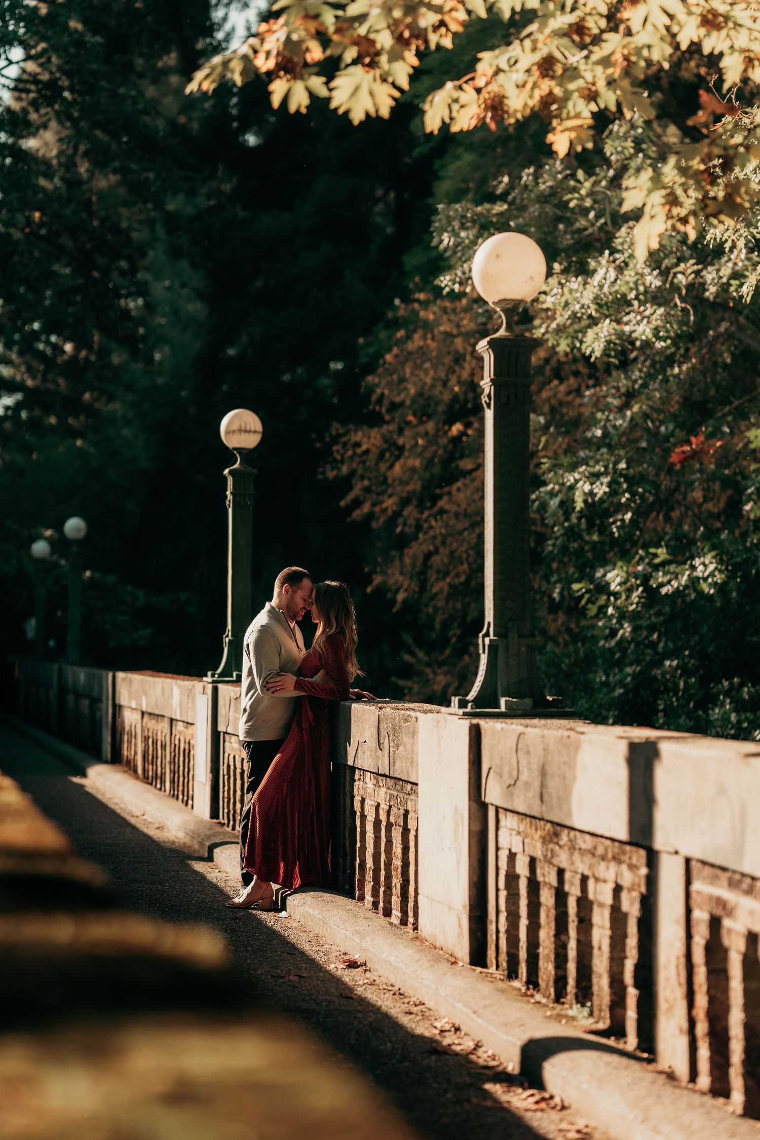 Couple sharing a kiss beside stone bridge railing under golden autumn foliage at sunset.