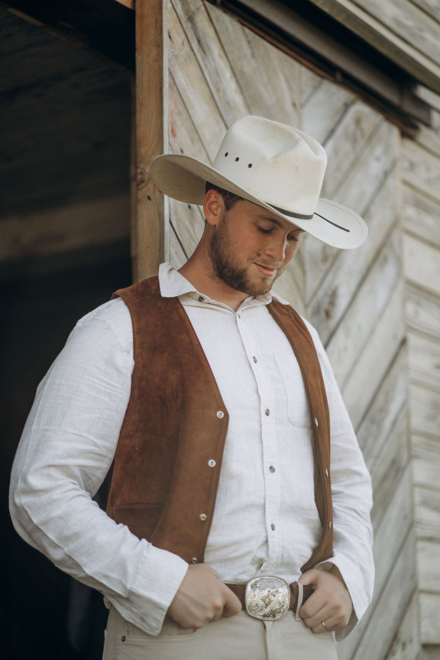 Groom in white shirt, suede vest, and cowboy hat looking down outside barn door.