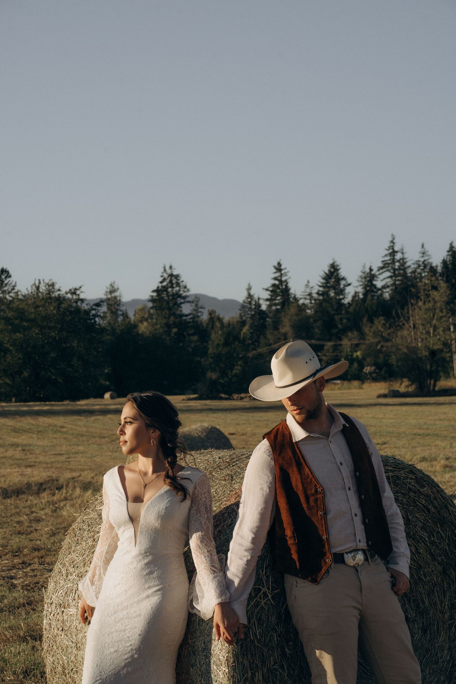 Bride and groom in western attire stand by a hay bale in a scenic field.