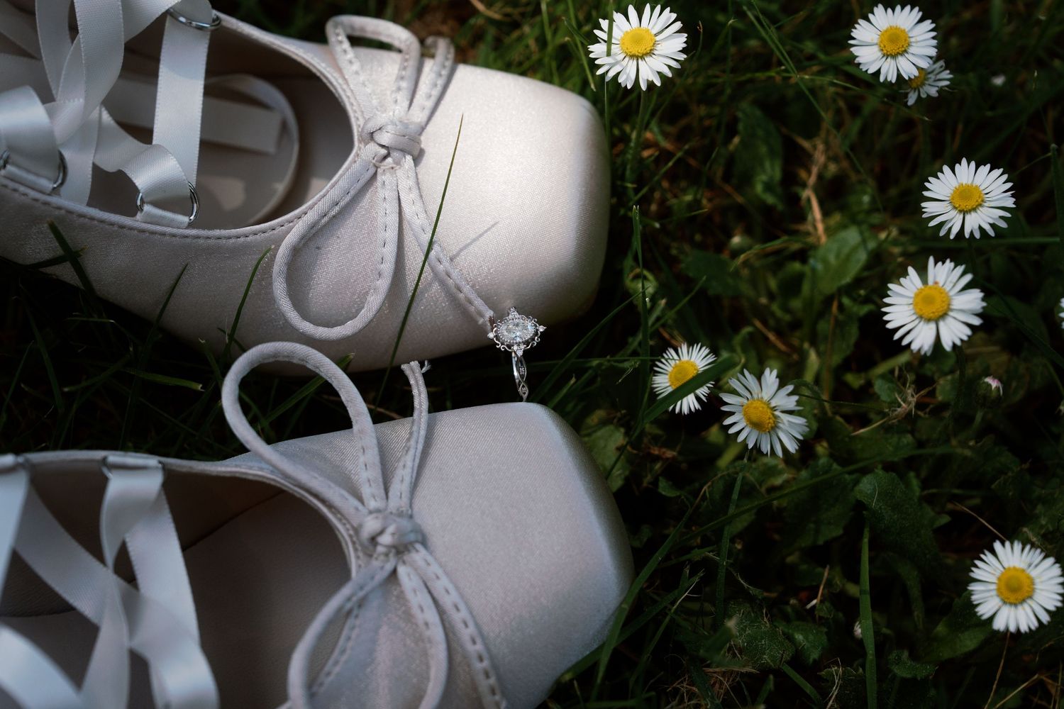 White bridal heels and delicate diamond ring rest among green grass and tiny white daisy wildflowers.