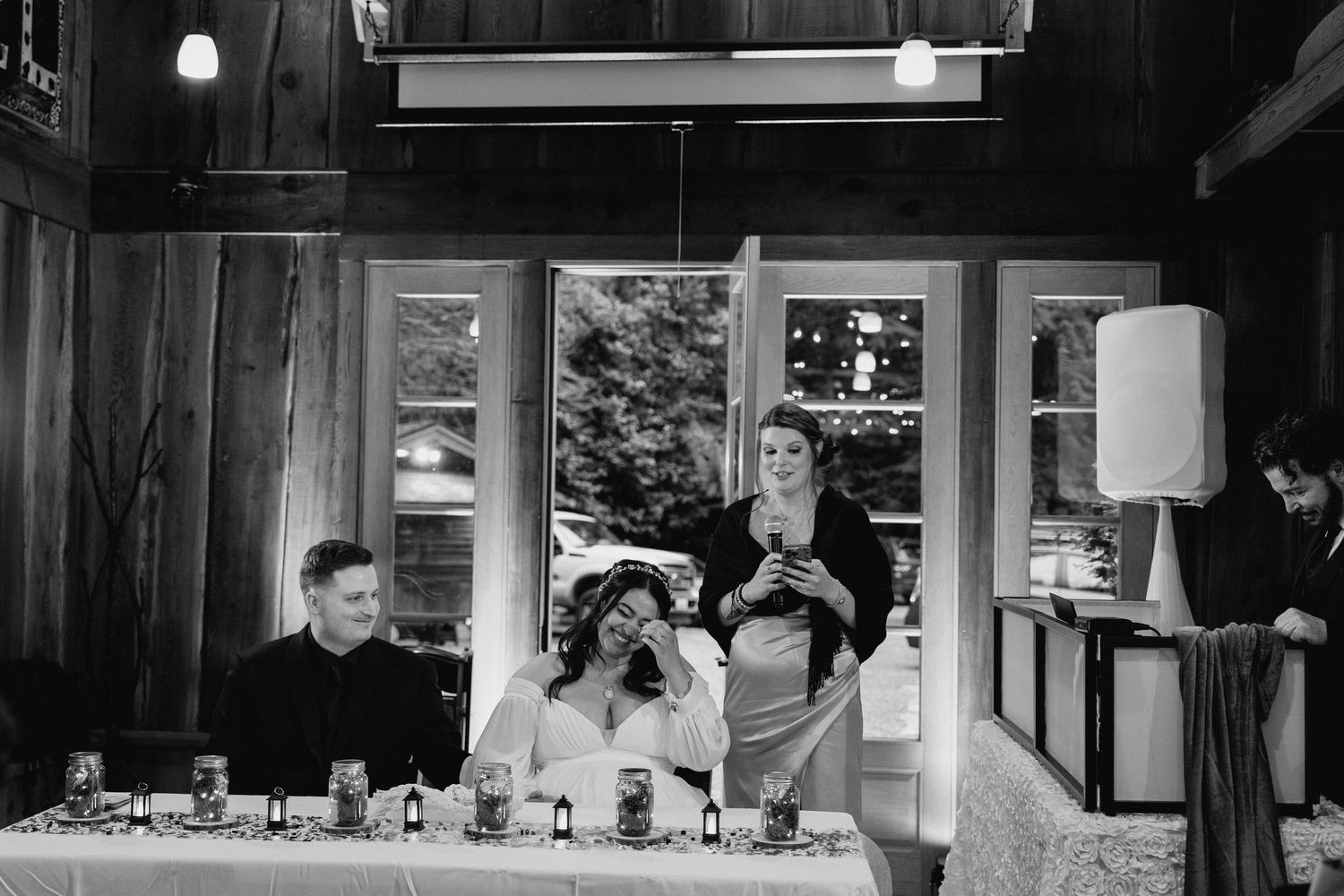 Bride laughs emotionally during wedding reception speech inside rustic venue in candid photo.
