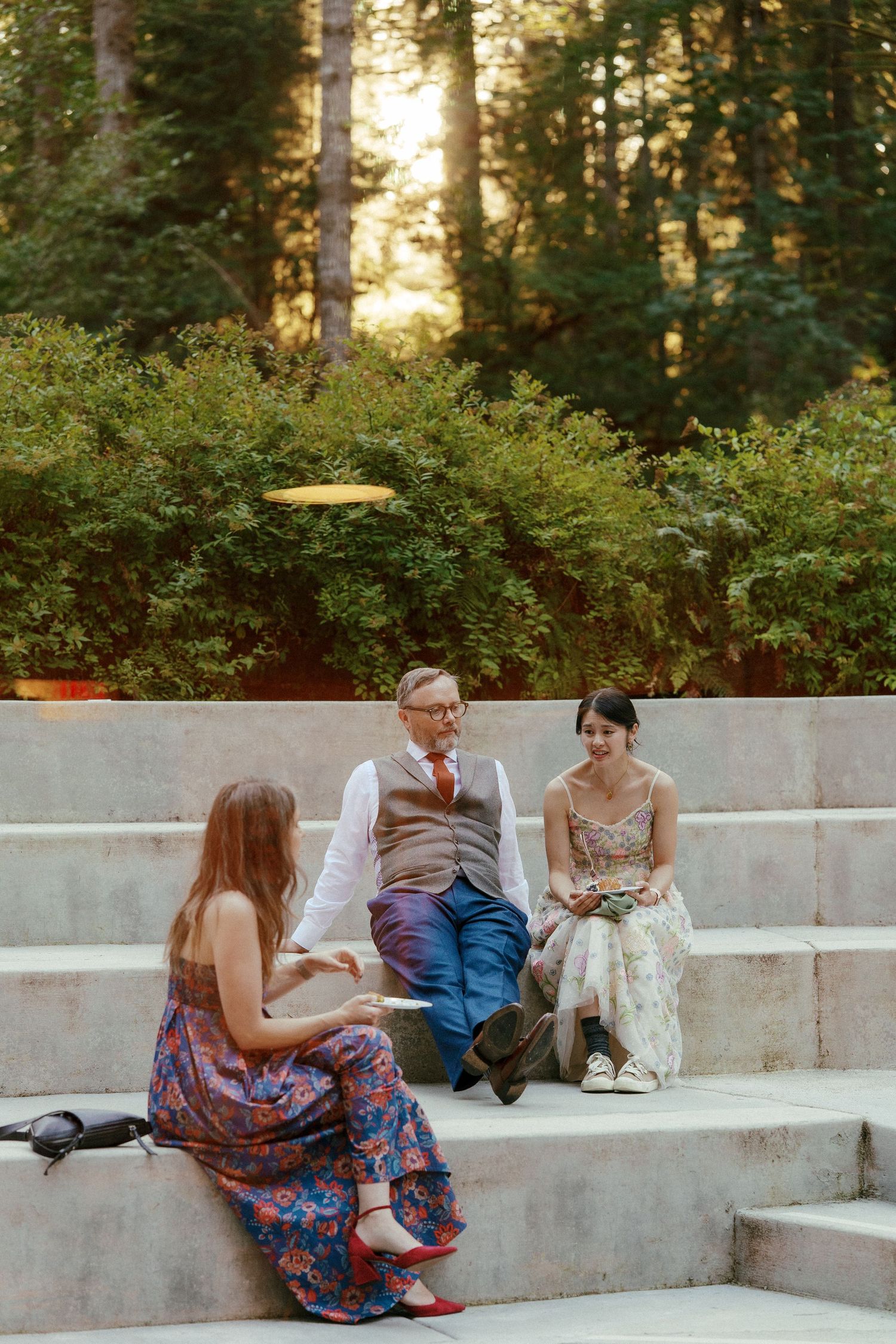 Three people conversing on stone steps surrounded by lush greenery at outdoor wedding venue.