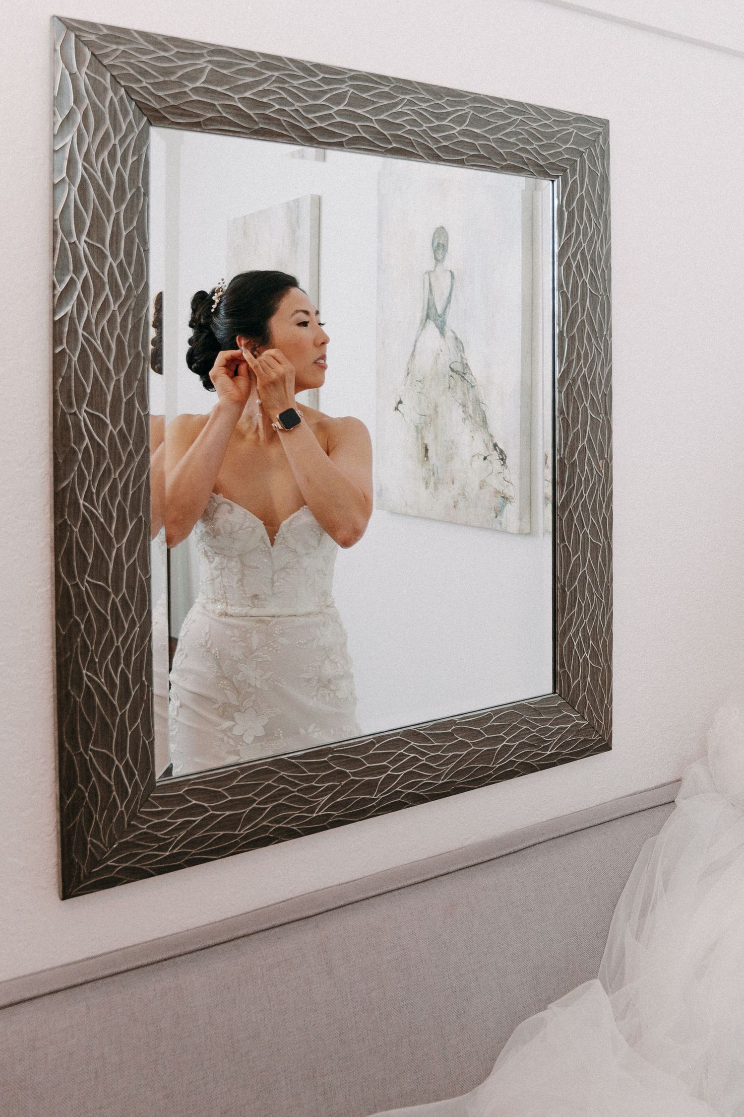 Bride in strapless lace wedding gown putting on earrings reflected in decorative framed mirror indoors.