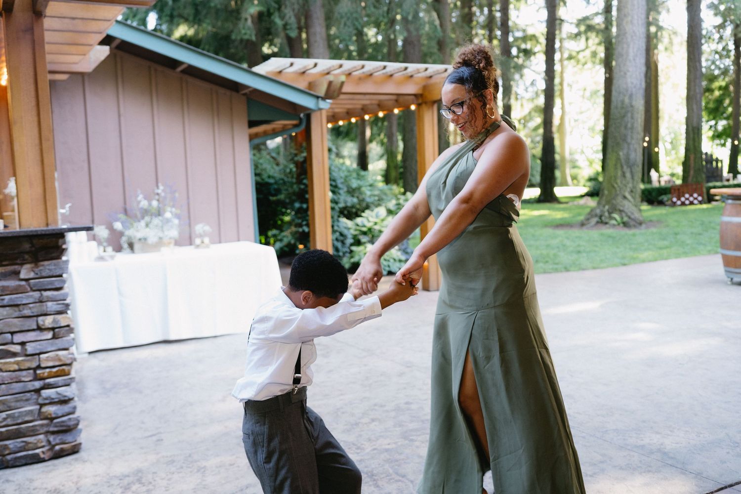 Woman in green dress dances playfully with young boy in suspenders at outdoor wedding reception.