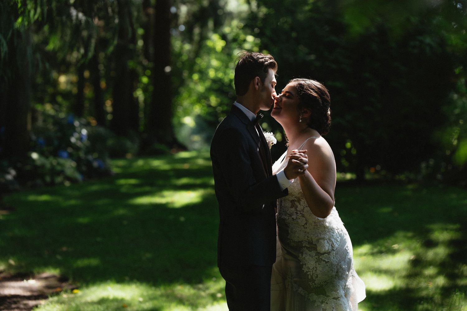 Bride and groom share a tender kiss in a shaded garden surrounded by tall lush trees.