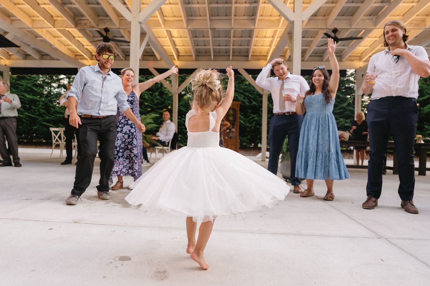 Flower girl in white tutu twirling barefoot surrounded by cheering guests at wedding reception.