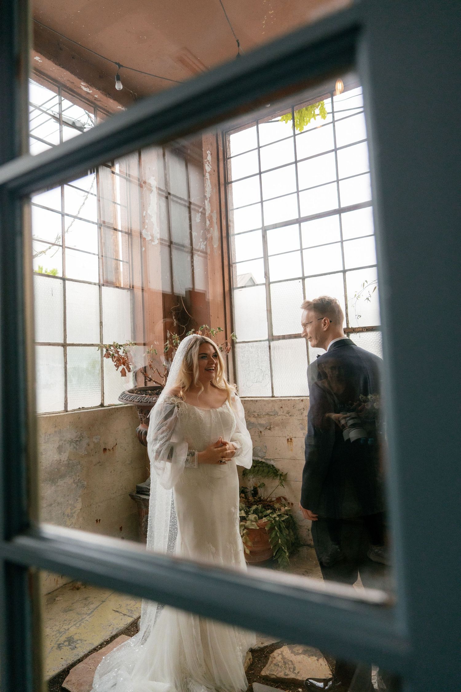 Bride in veil and groom viewed through blue window panes in sunlit industrial wedding venue.