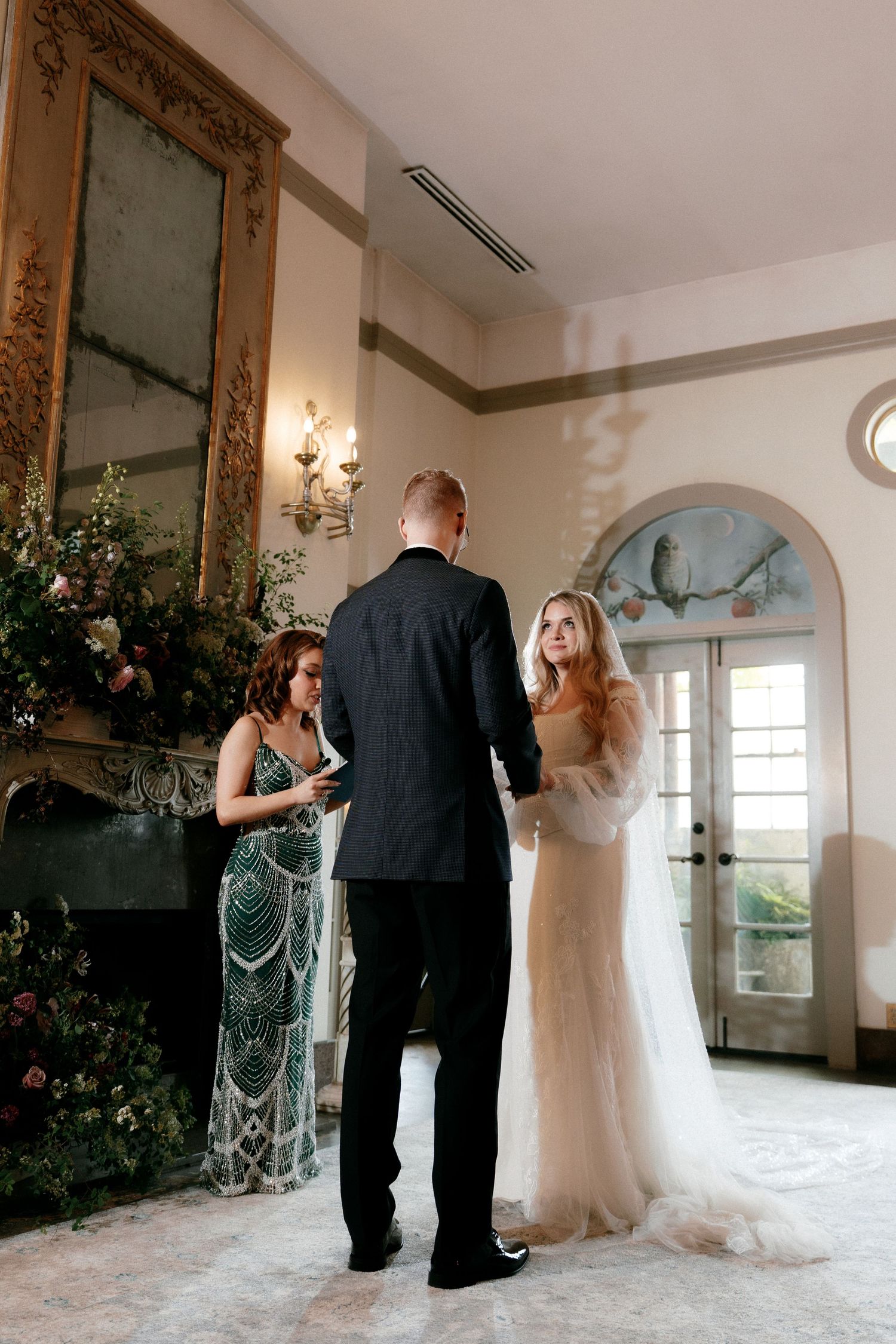 Bride and groom exchange vows at an ornate fireplace altar as an officiant reads during their indoor wedding.