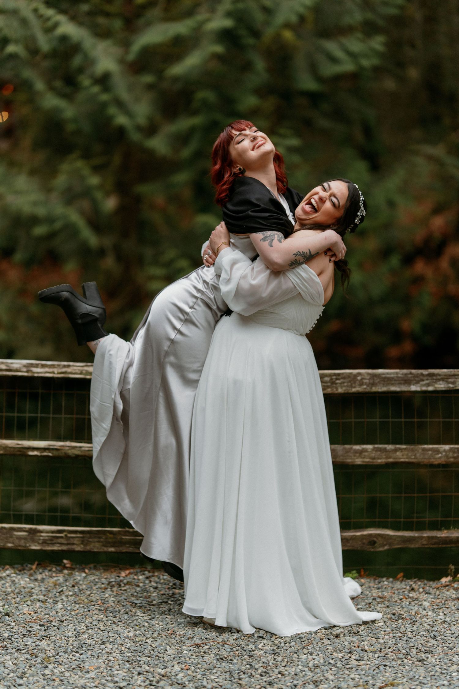 Two brides laughing joyfully as one lifts the other at outdoor wedding.