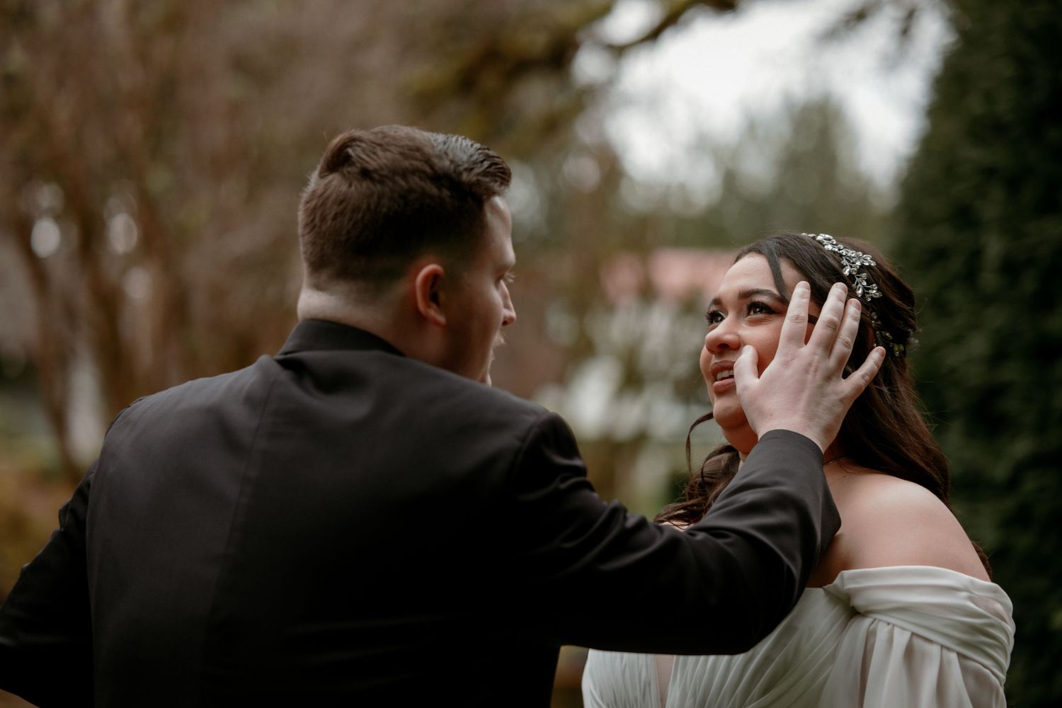 Groom tenderly touching bride's face as she smiles, standing outdoors in a lush garden setting.
