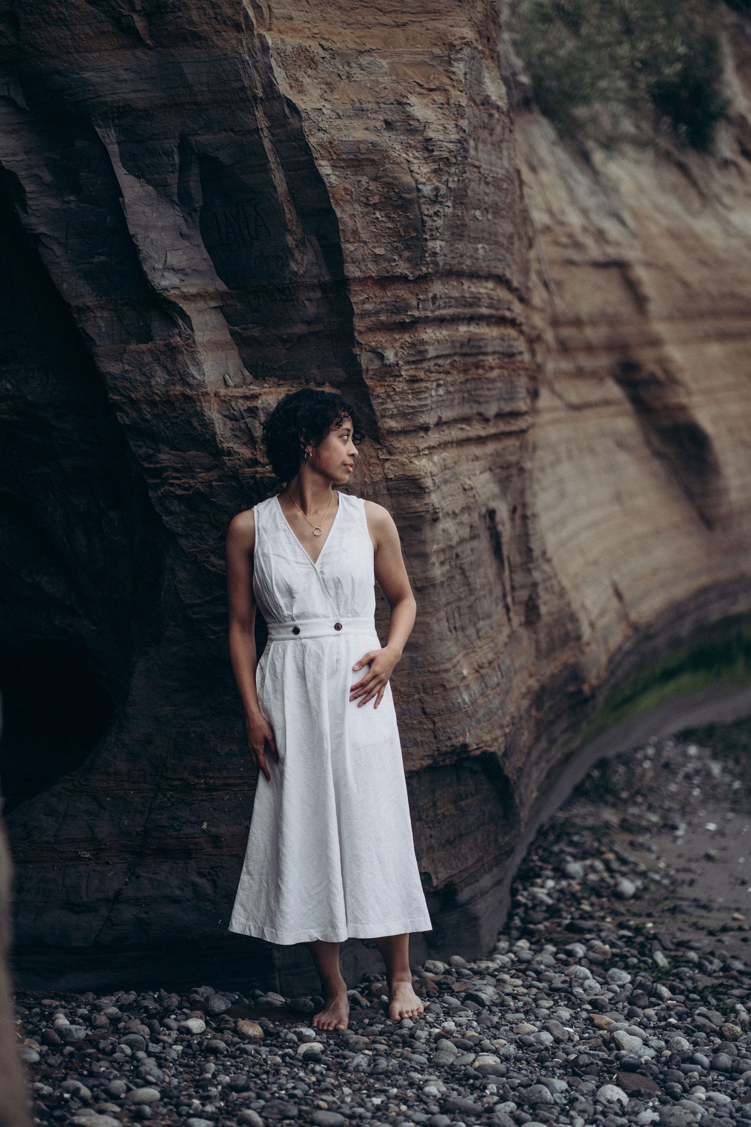Barefoot woman in white linen dress stands against dramatic layered sandstone cliff wall.