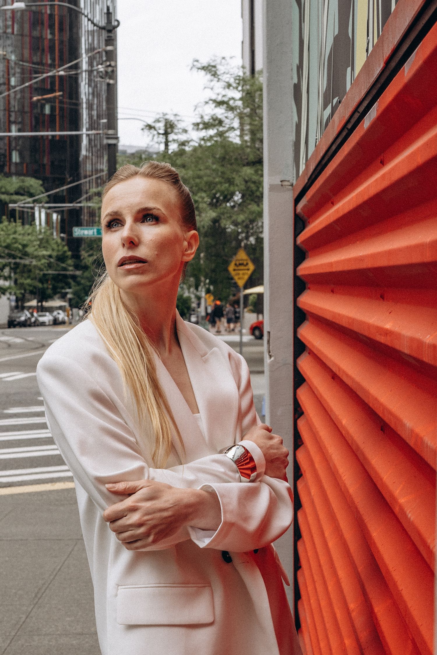 Blonde woman in white blazer with arms crossed leans against orange corrugated wall on urban NYC street.
