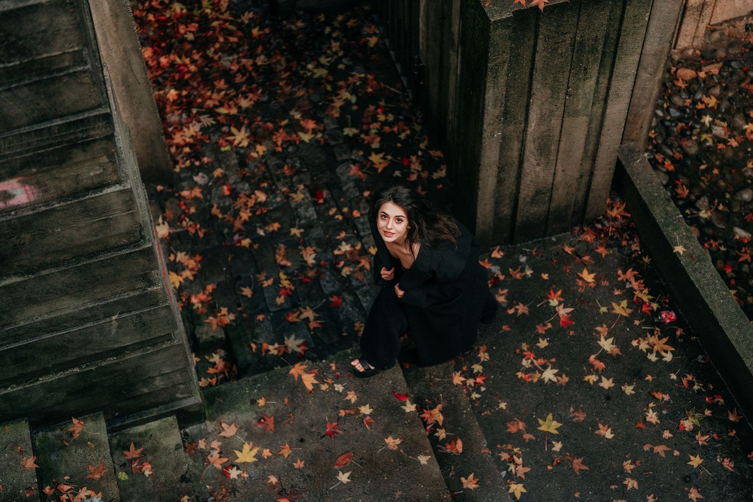 Young woman in black outfit sits on autumn leaf-covered wooden steps viewed from above.
