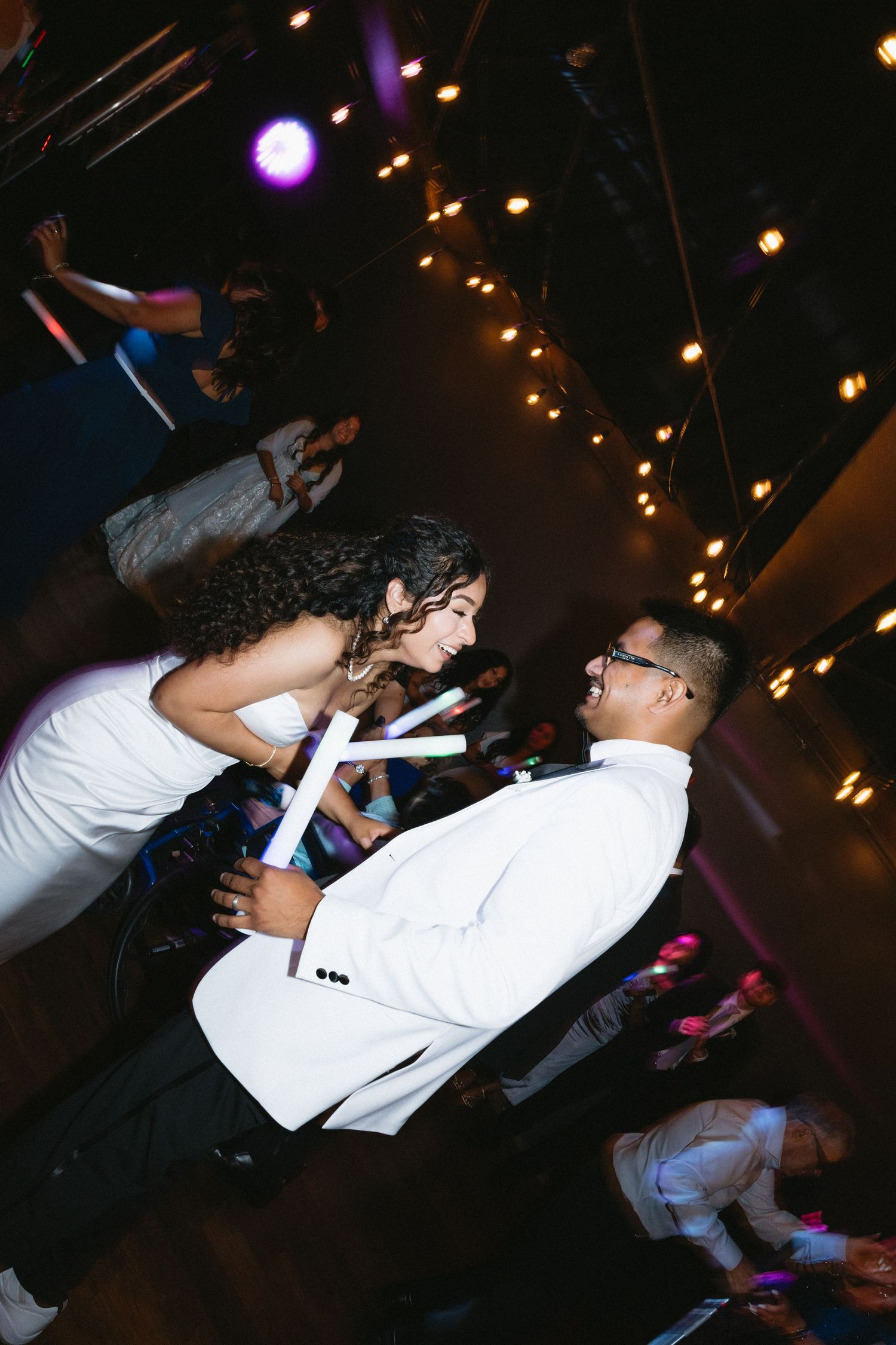 Bride and groom dance together with glow sticks surrounded by guests at a festive wedding reception.