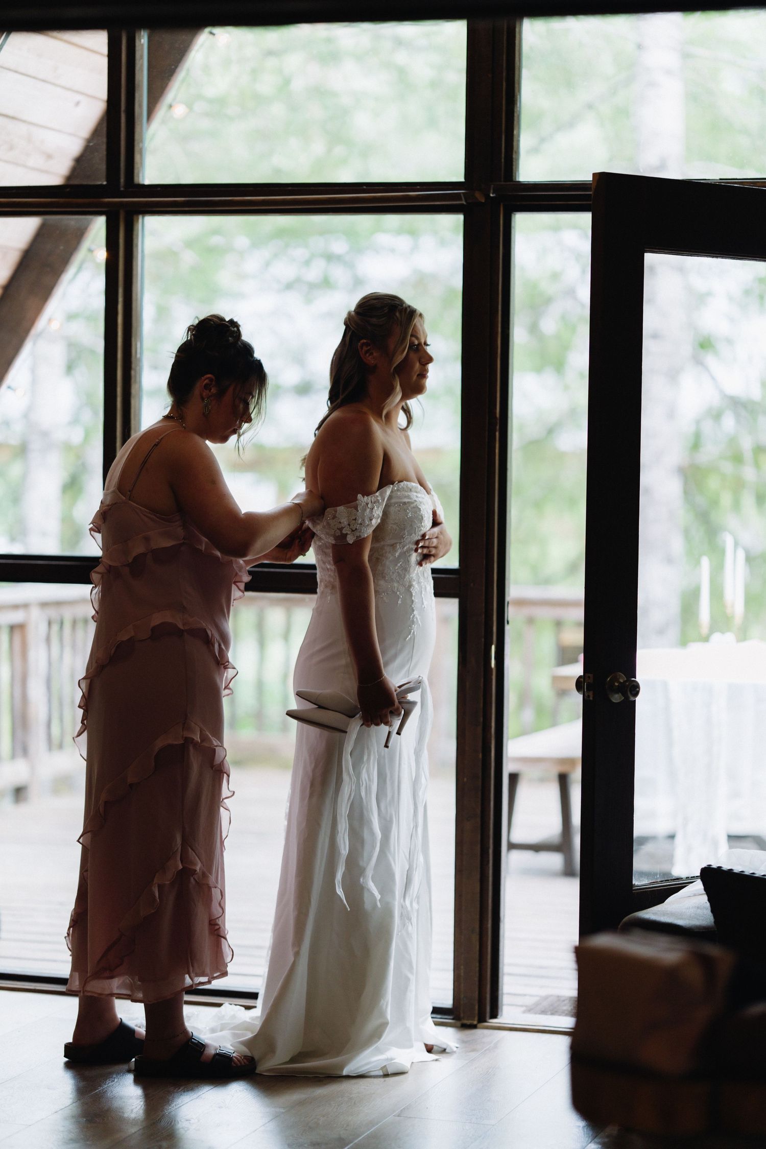 Bridesmaid helping bride with her off-shoulder wedding dress near large windows before the ceremony.