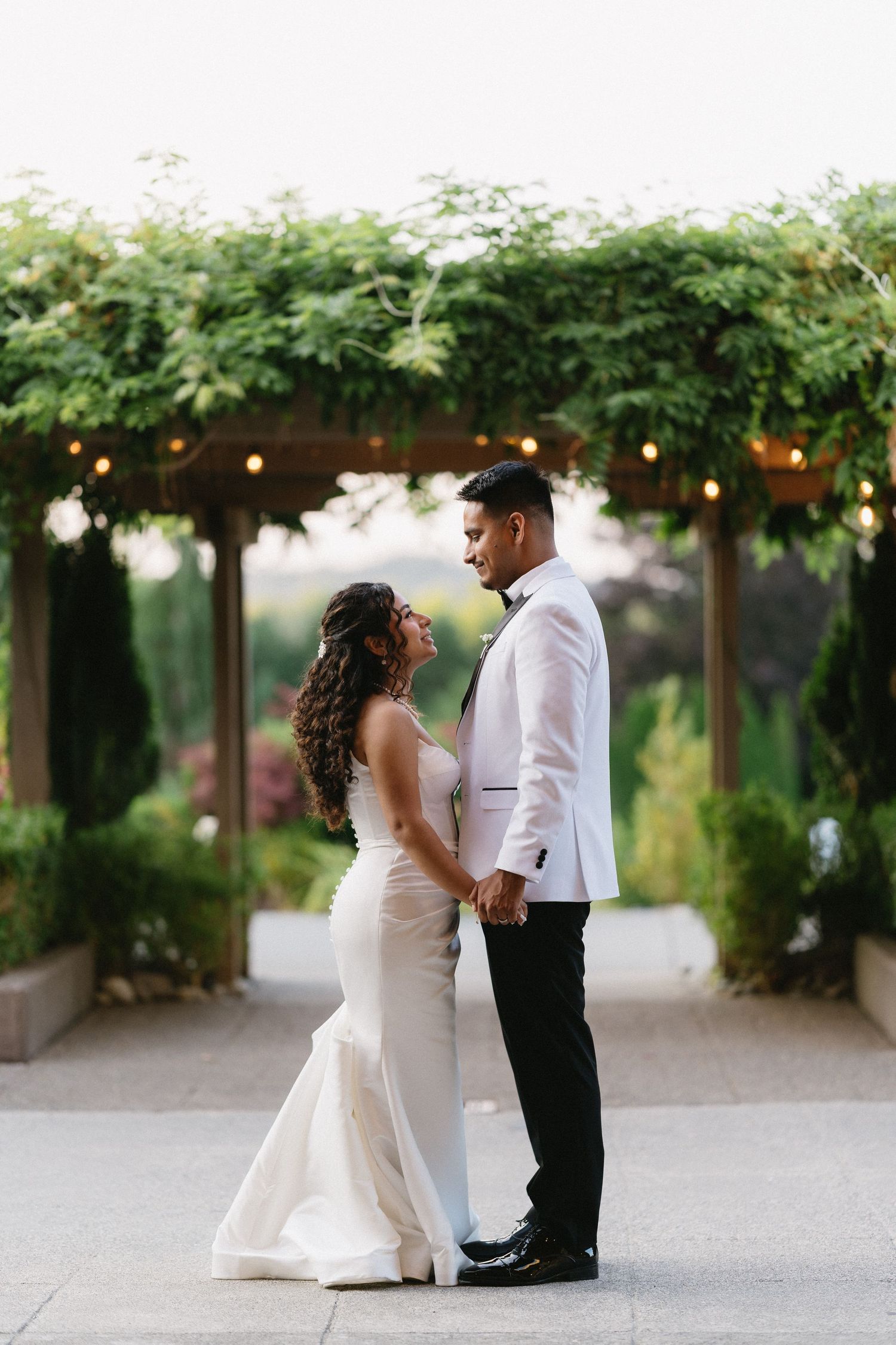 Bride and groom hold hands and gaze at each other under a lush green pergola at sunset.
