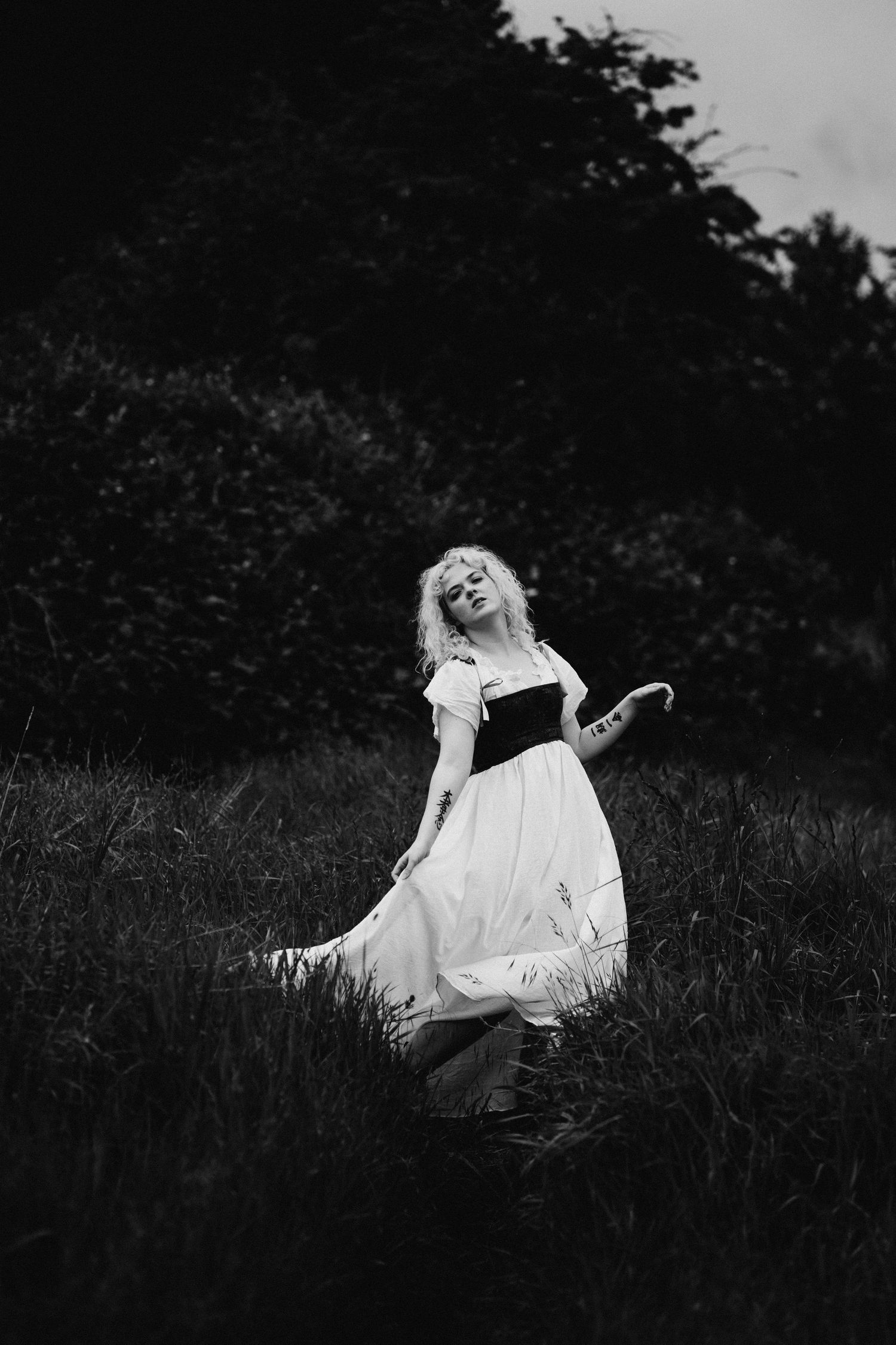 A blonde woman in a white dress and black corset poses in a dark grassy field in a dramatic black and white photo.