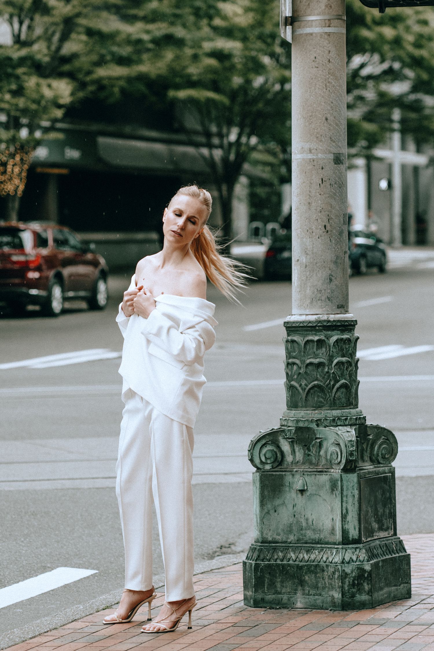 Blonde woman in white off-shoulder blazer and pants stands by ornate street lamp at city crosswalk.