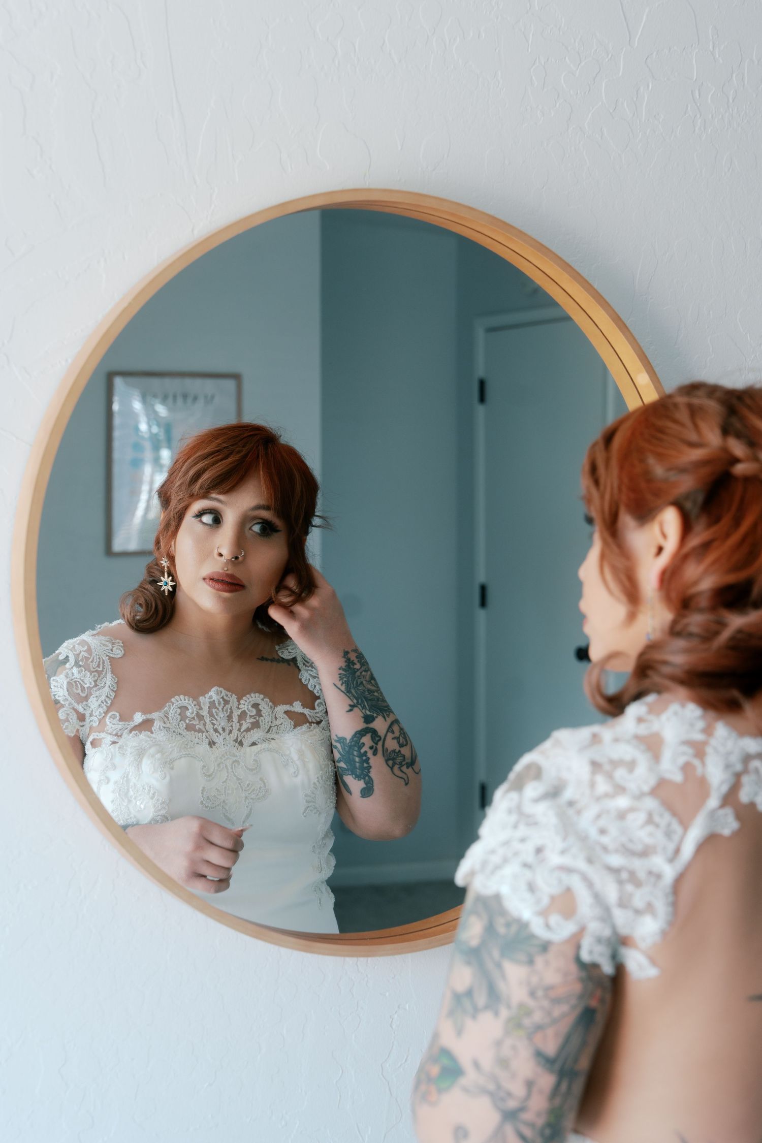 Tattooed redhead bride in lace wedding gown adjusts earring while gazing at her reflection in round mirror.