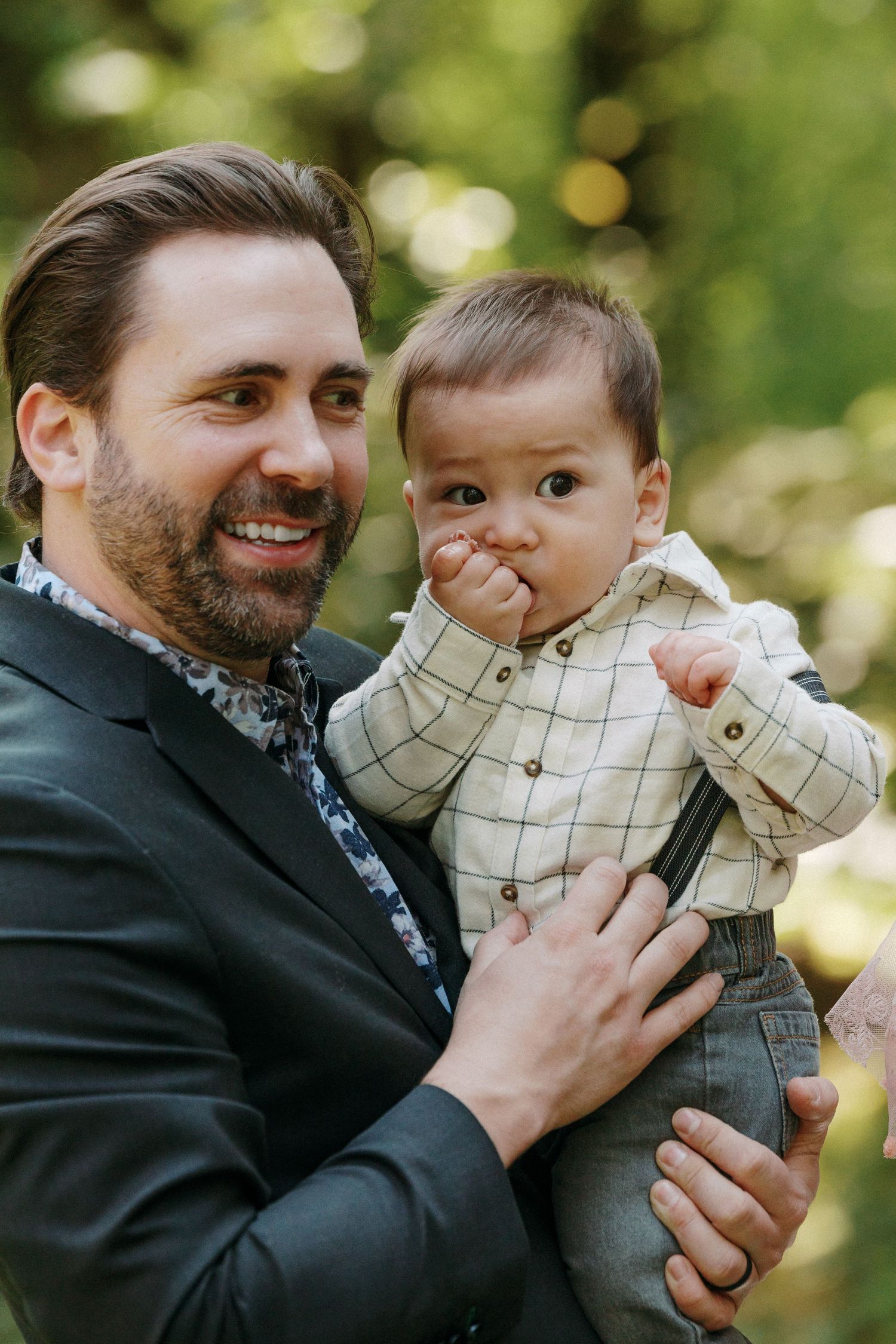 Smiling bearded man in dark suit holding adorable baby in plaid shirt at outdoor wedding event.