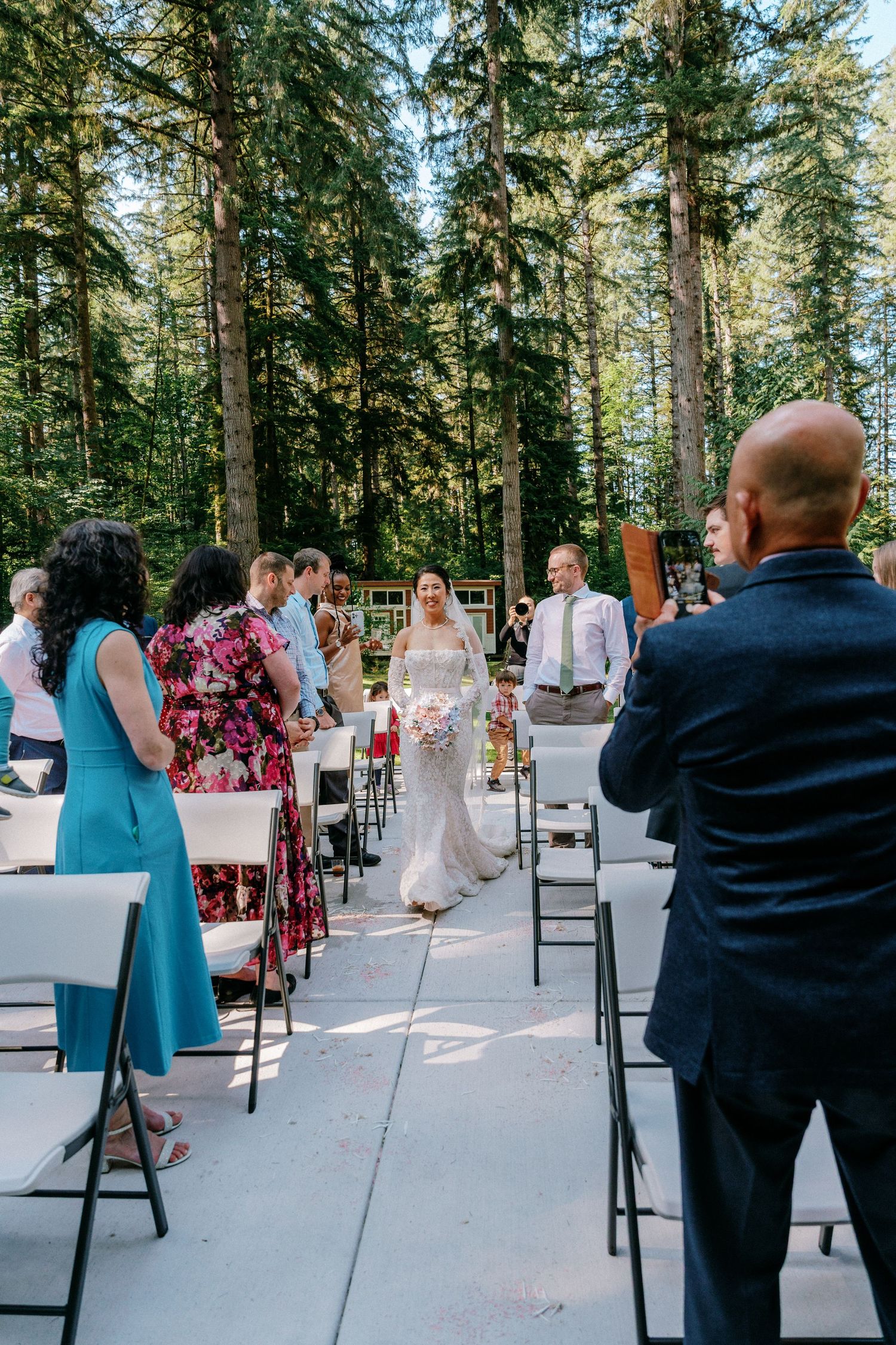 Bride walks down outdoor wedding aisle surrounded by tall evergreen trees as guests stand and watch.
