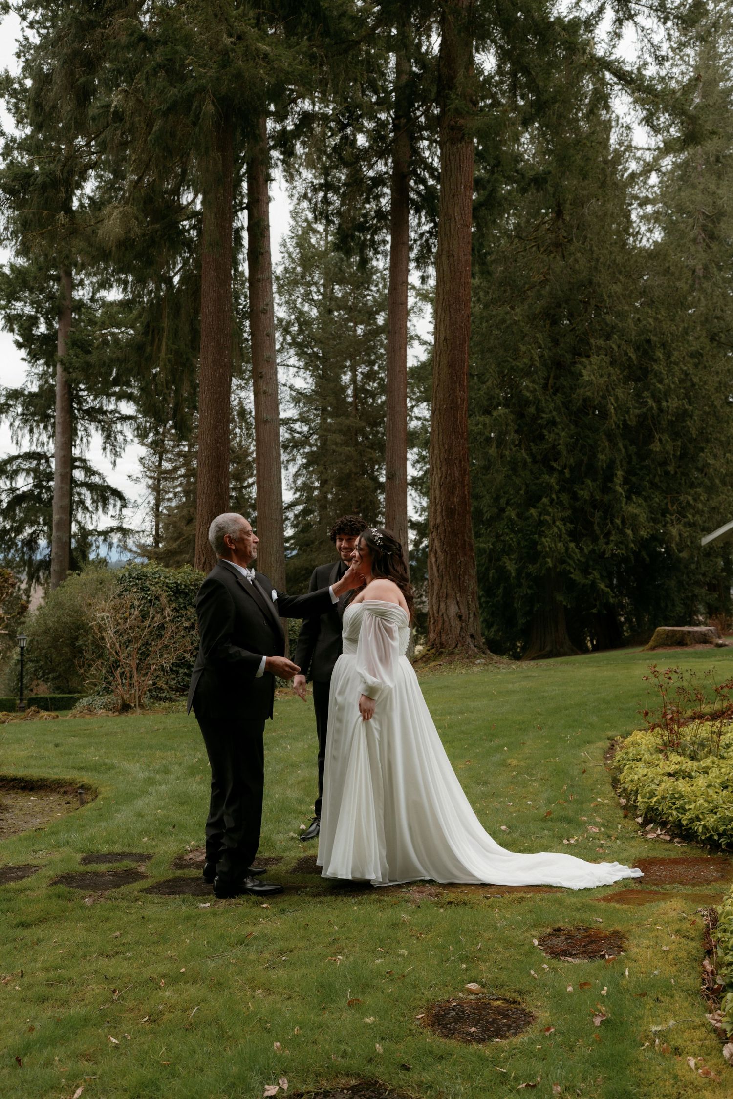 Bride in flowing white off-shoulder gown holds hands with elderly man during outdoor forest wedding ceremony.