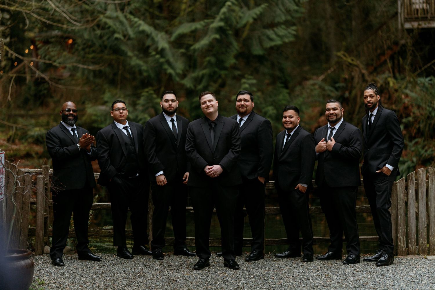 Group of eight groomsmen in black suits posing at outdoor forest wedding.