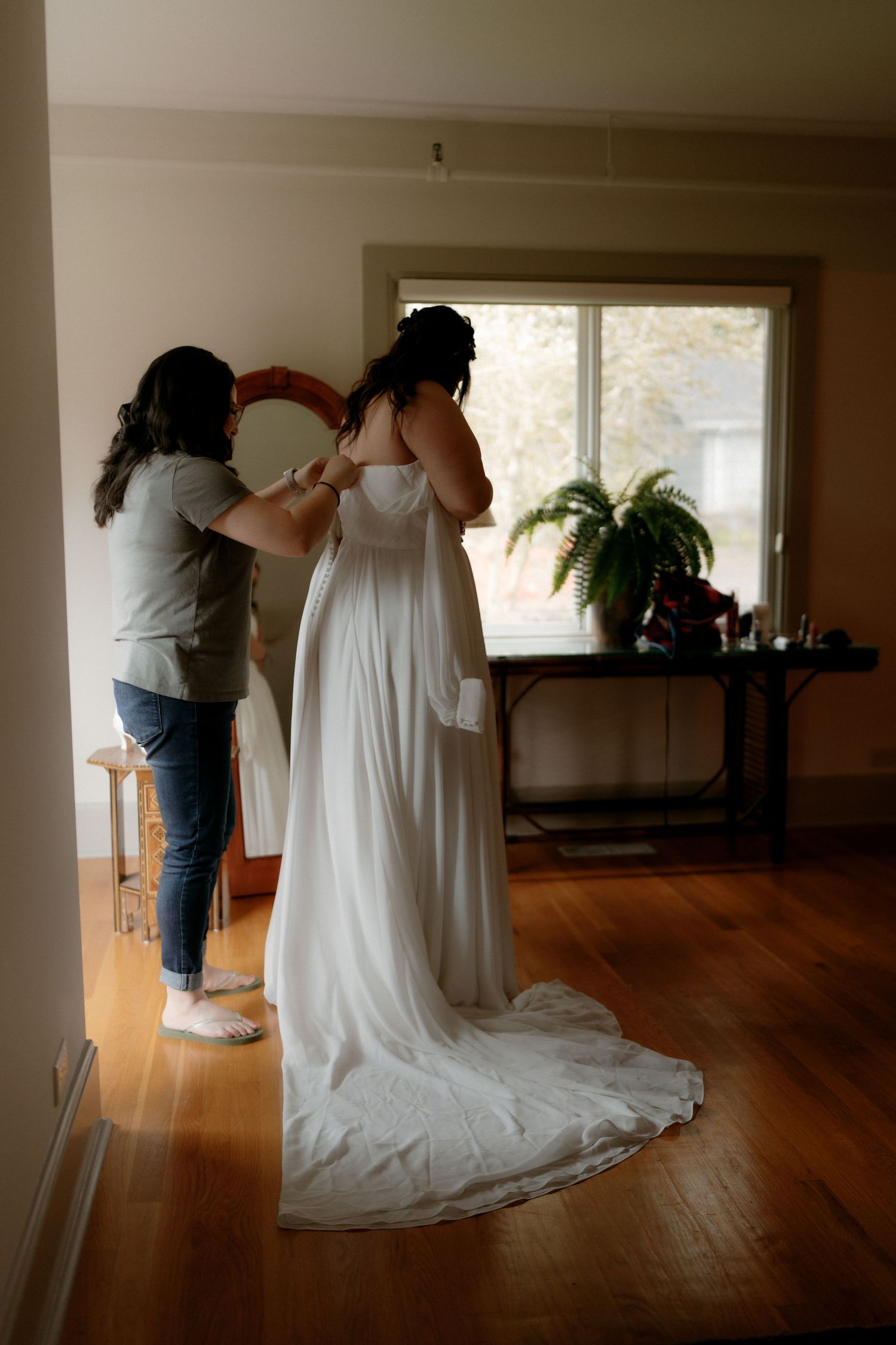 Bride being helped into her white wedding gown by assistant near a sunlit window before ceremony.