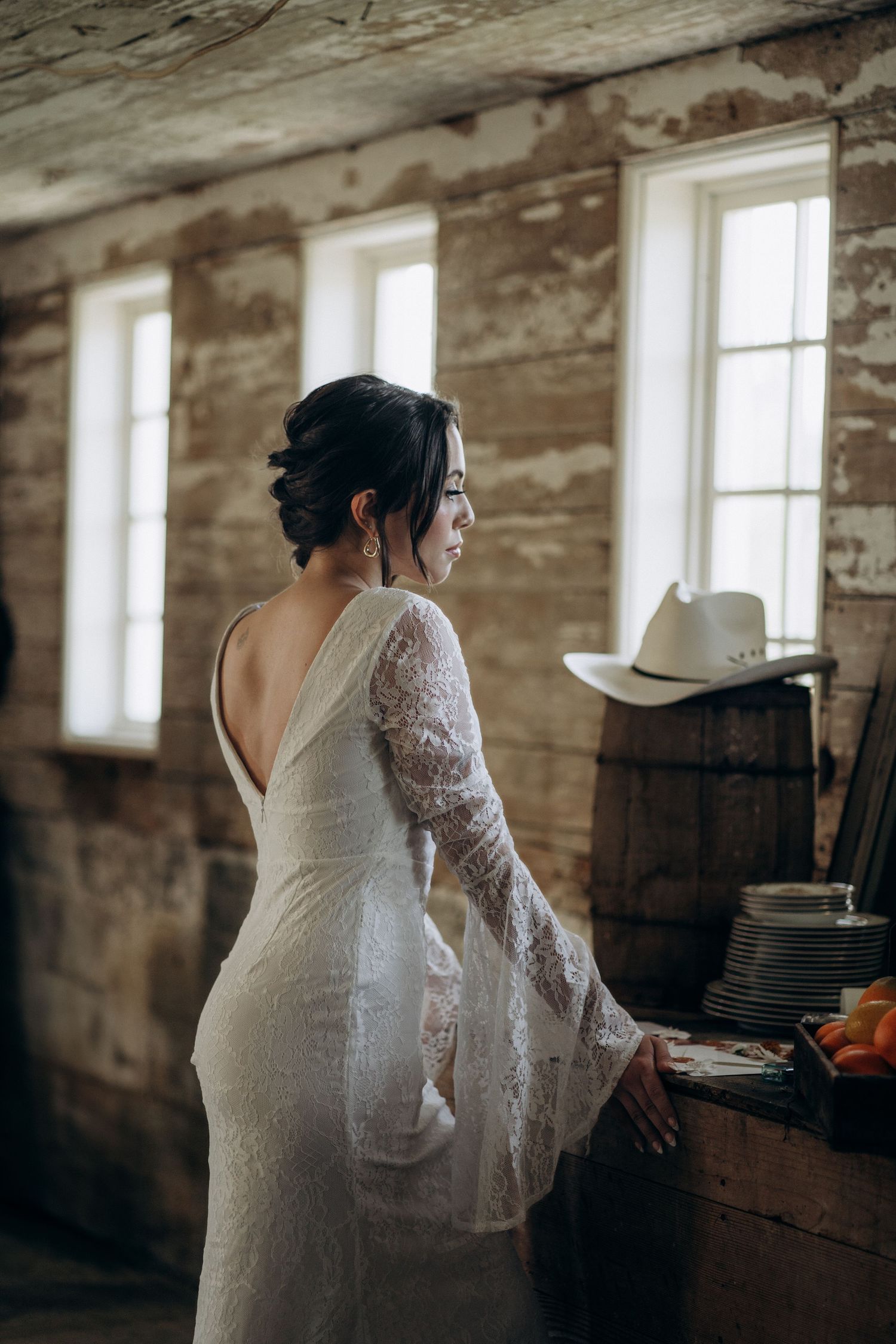 Bride in backless white lace bell-sleeve wedding dress standing by windows in rustic stone barn.