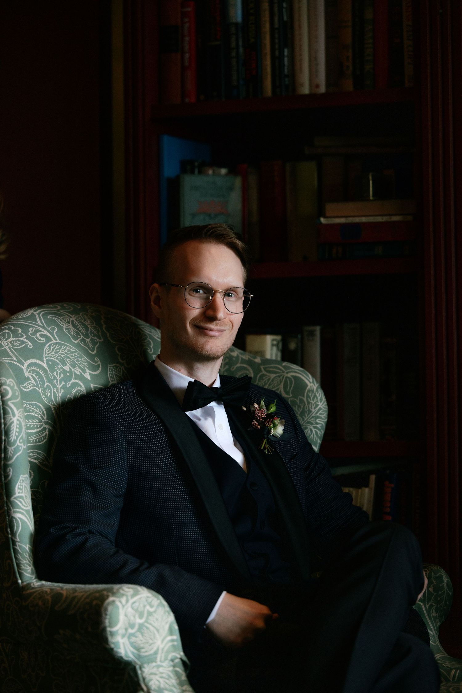 Smiling groom in black tuxedo with boutonniere seated in an armchair beside a wooden bookshelf.
