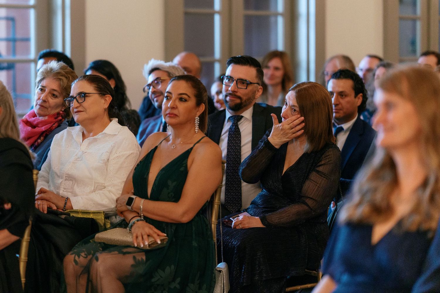 Emotional wedding guests in formal attire watching the ceremony, with one woman touching her face in tears.