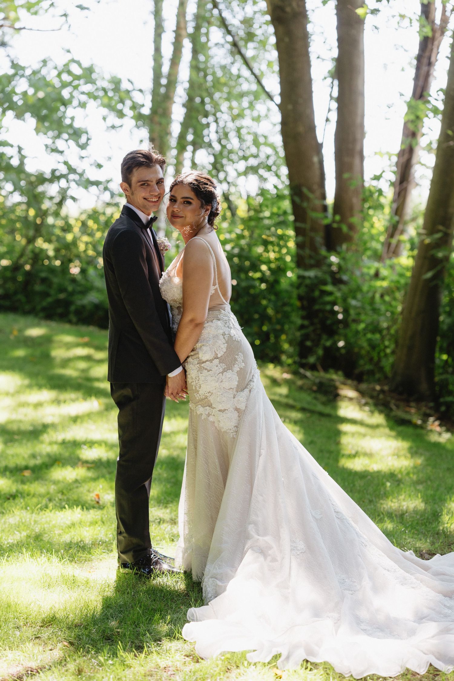 Bride and groom pose together smiling in a sunlit garden, she in a lace wedding gown with train, he in a black suit.