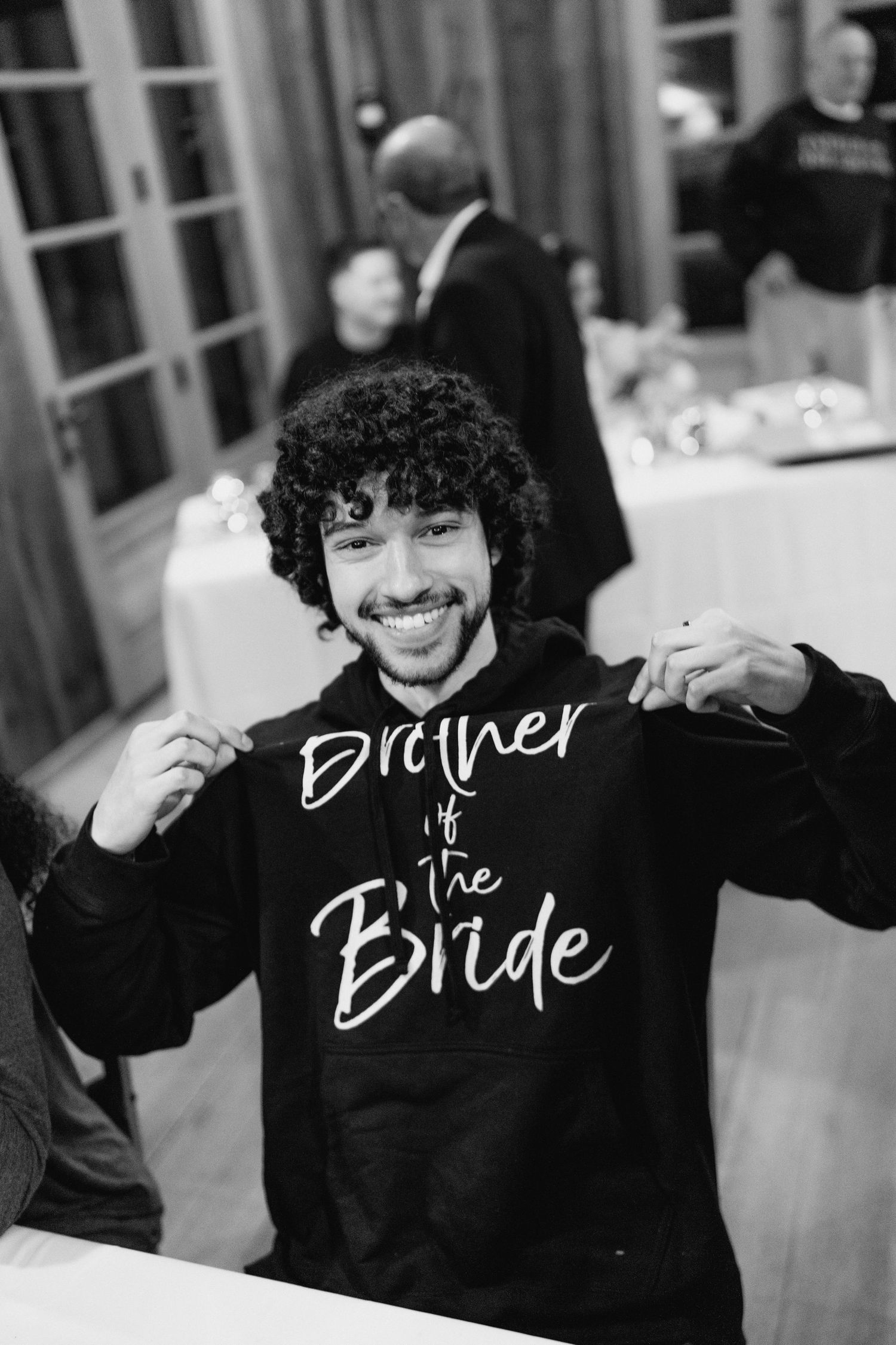 Young man smiling and holding up a black Brother of the Bride hoodie at a wedding celebration.
