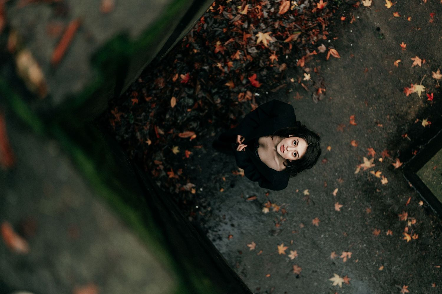 Aerial view of a person in black coat surrounded by fallen autumn leaves on pavement.