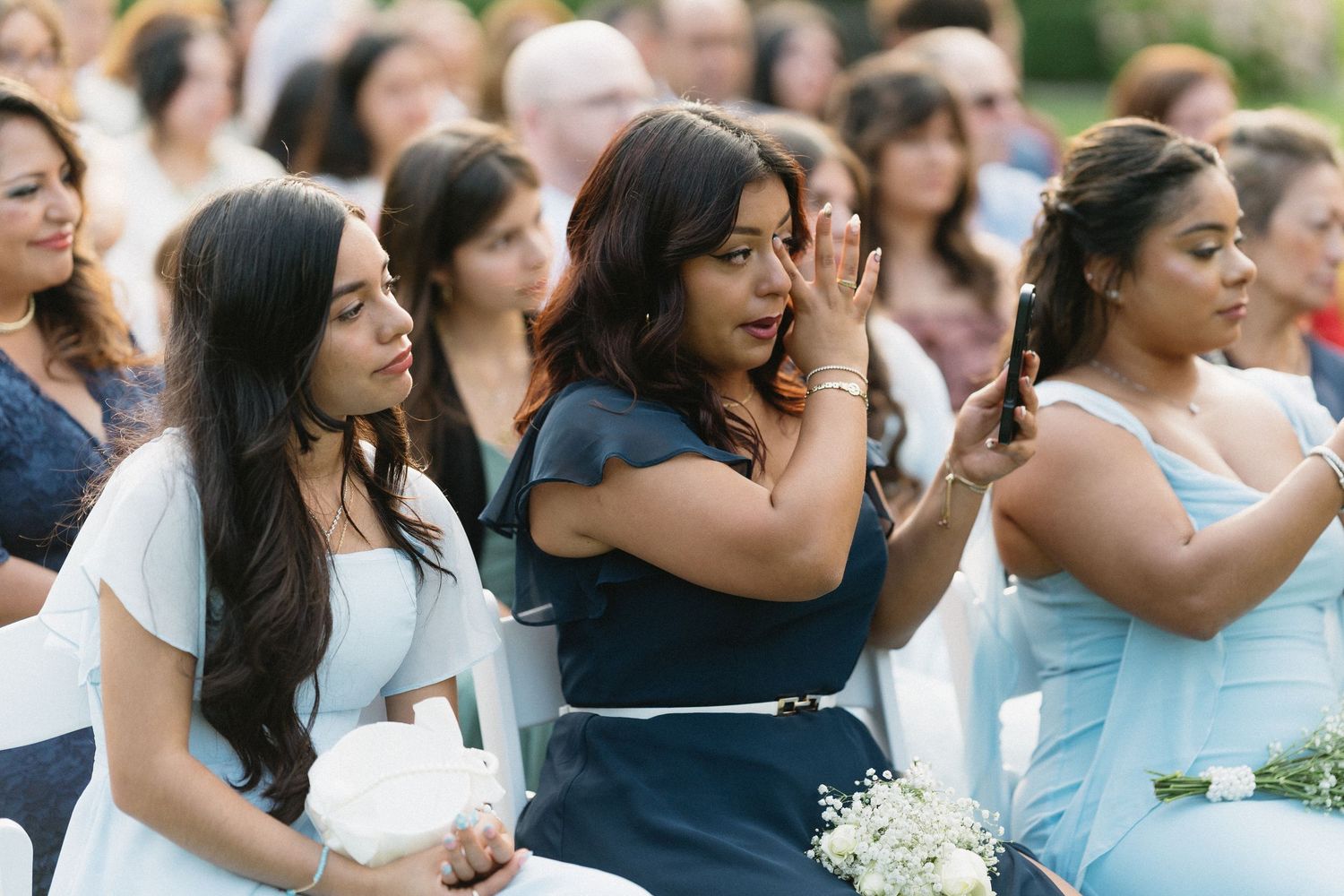 Emotional wedding guest in navy dress reacts with tears during outdoor ceremony.