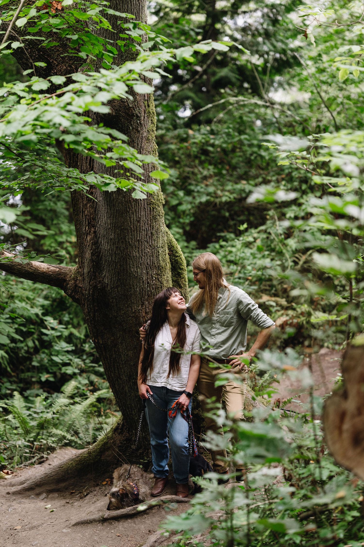 A couple shares a tender moment beside a large mossy tree on a forest trail.