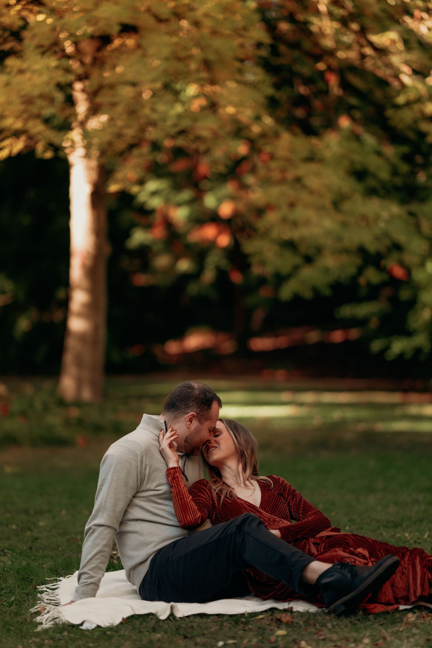 Couple kissing while seated on a white blanket in a sunlit autumn park setting.