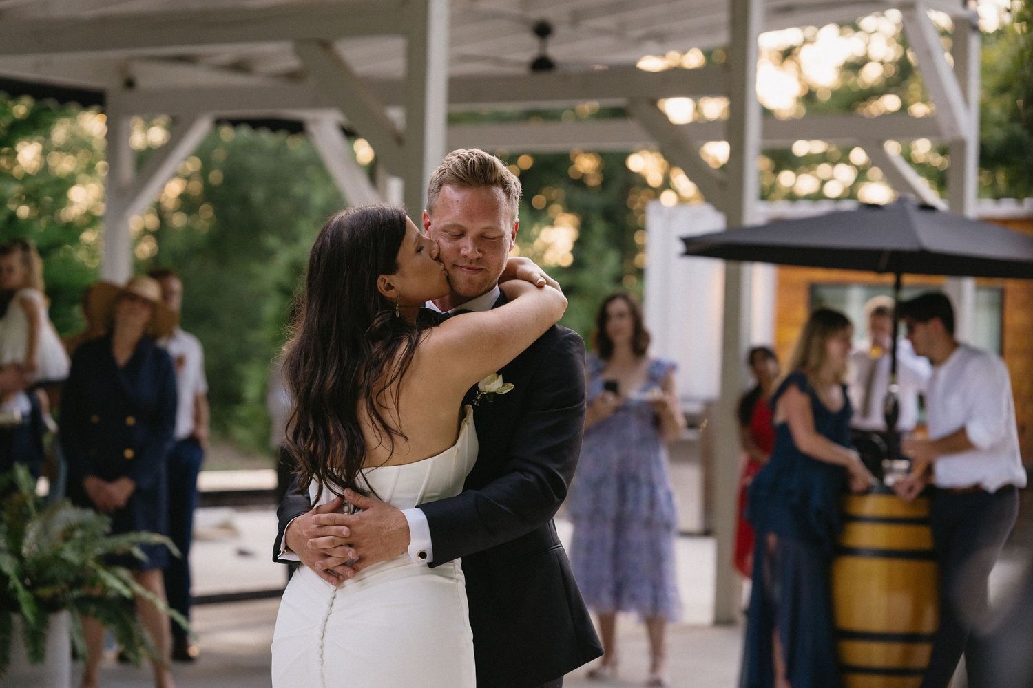 Bride and groom share their first dance kiss on an outdoor patio with string lights and guests in the background.