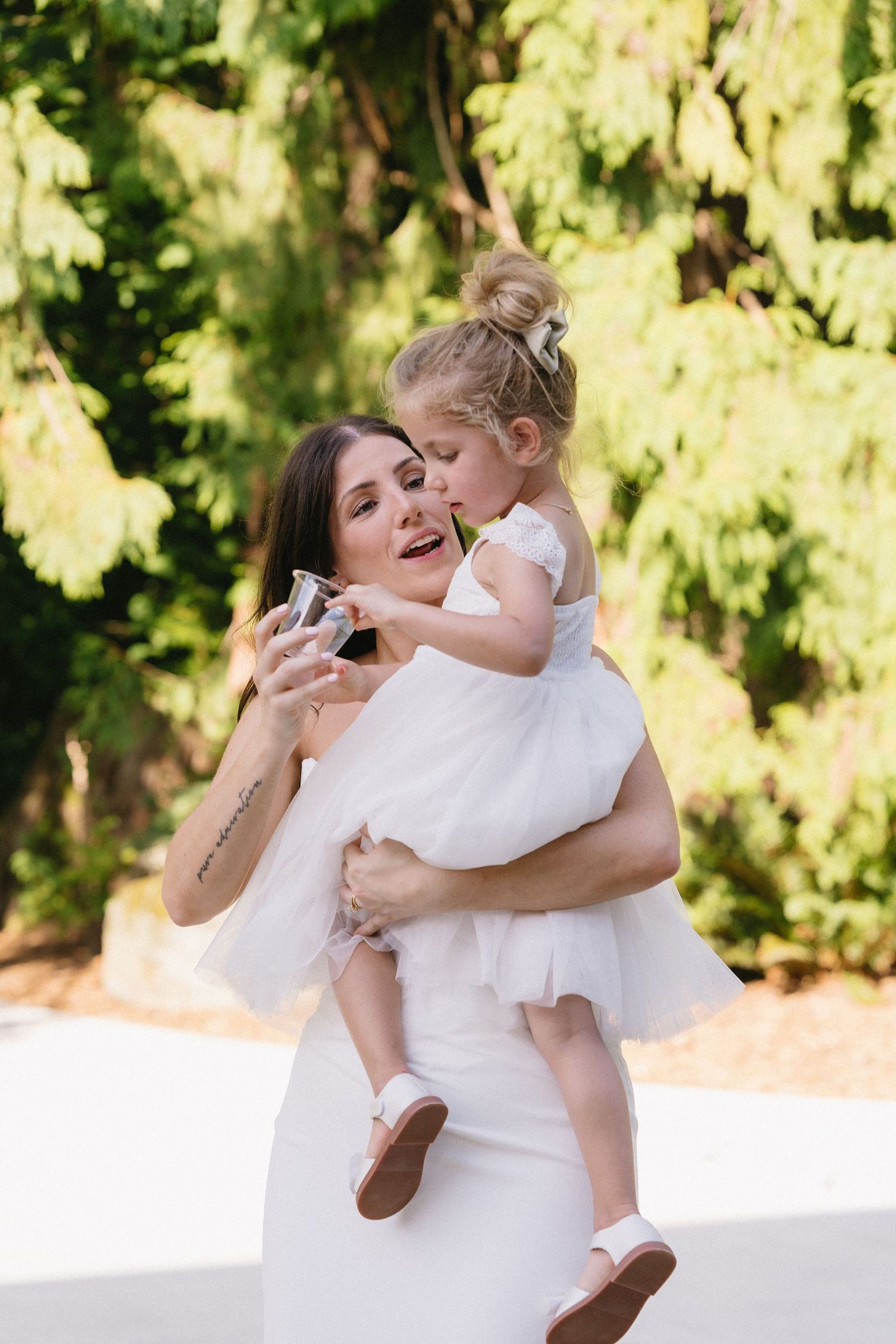 Bride in white gown holding a young flower girl who kisses her cheek outdoors.