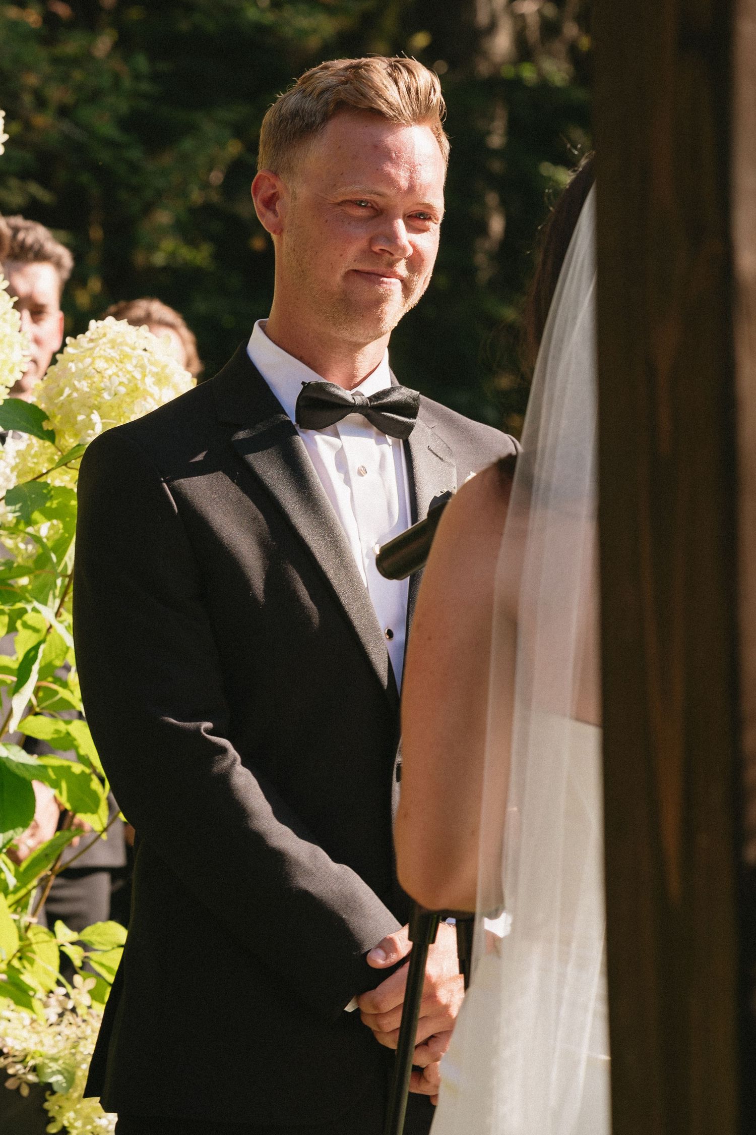 Groom in black tuxedo smiling lovingly at bride during outdoor wedding ceremony.