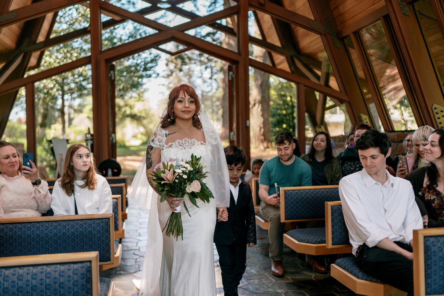 Bride in lace gown walking down chapel aisle with young boy escort past seated guests.