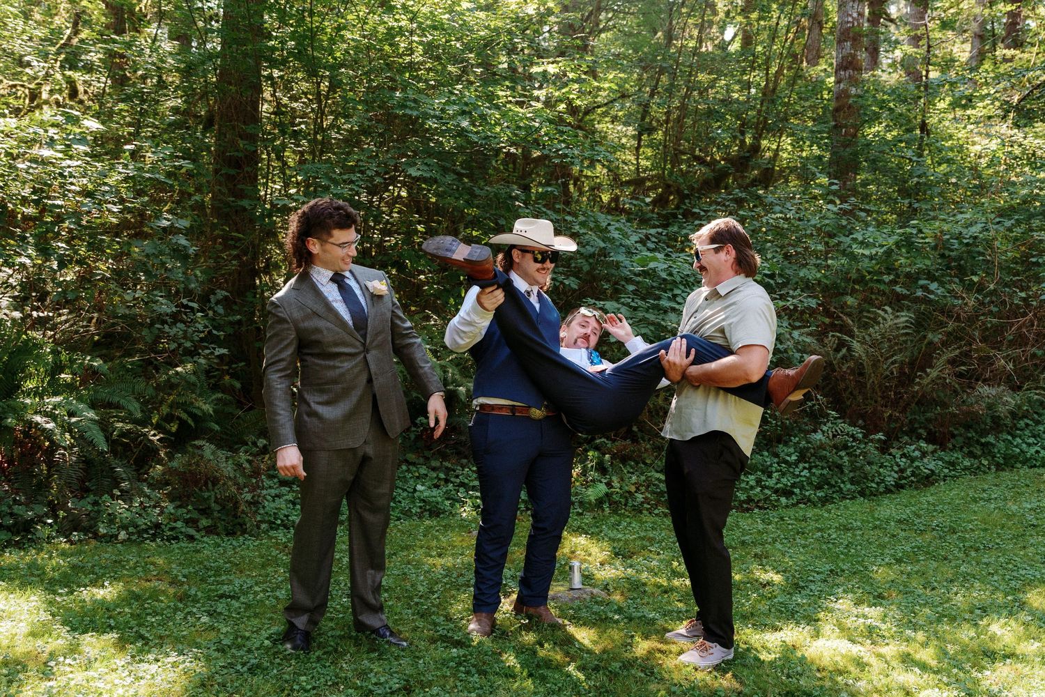 Three wedding guests pose playfully outdoors, with one man carrying another while a third stands nearby in formal attire.
