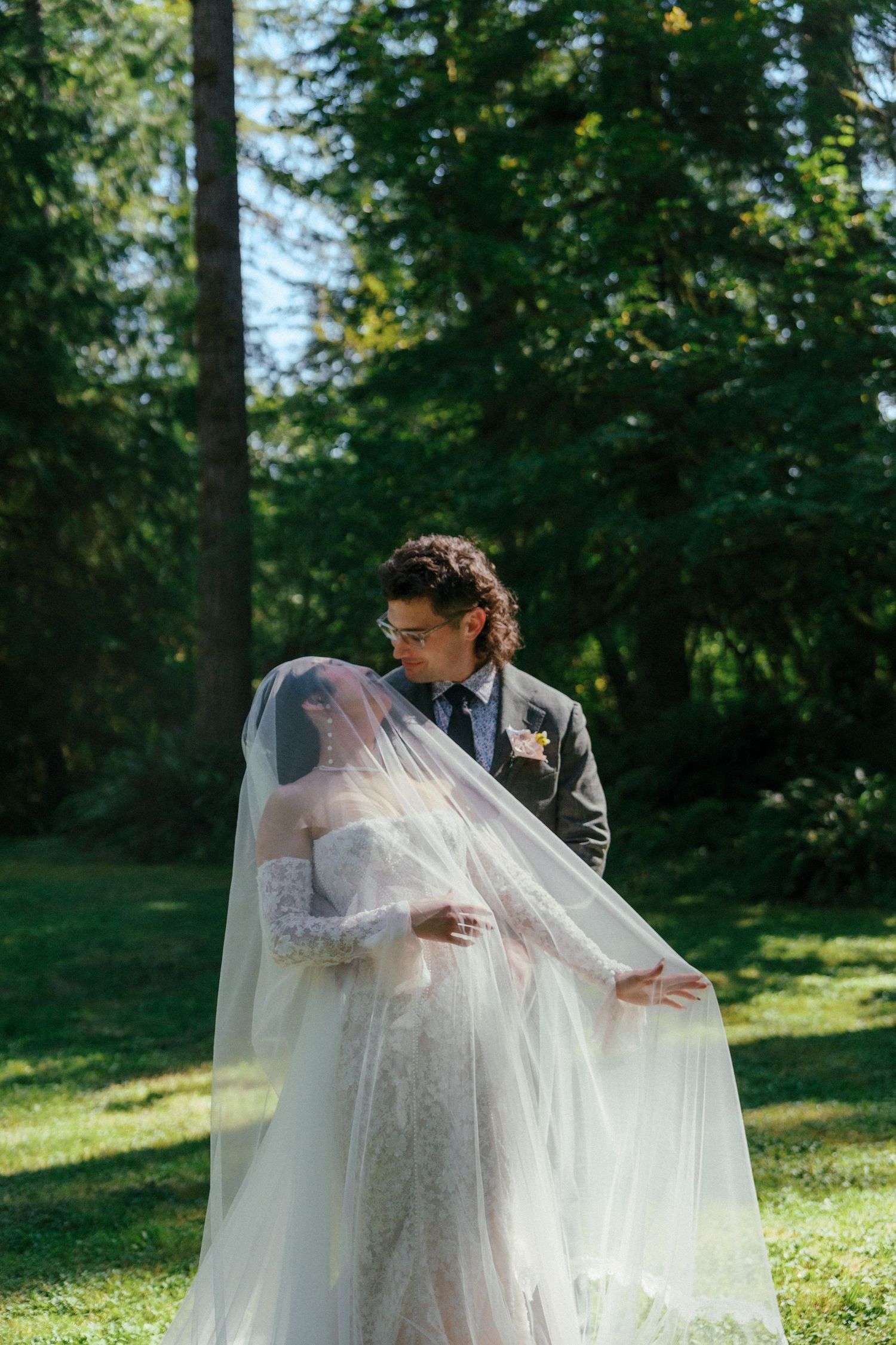 Groom lifts bride's cathedral veil as couple embraces in lush green forest setting.