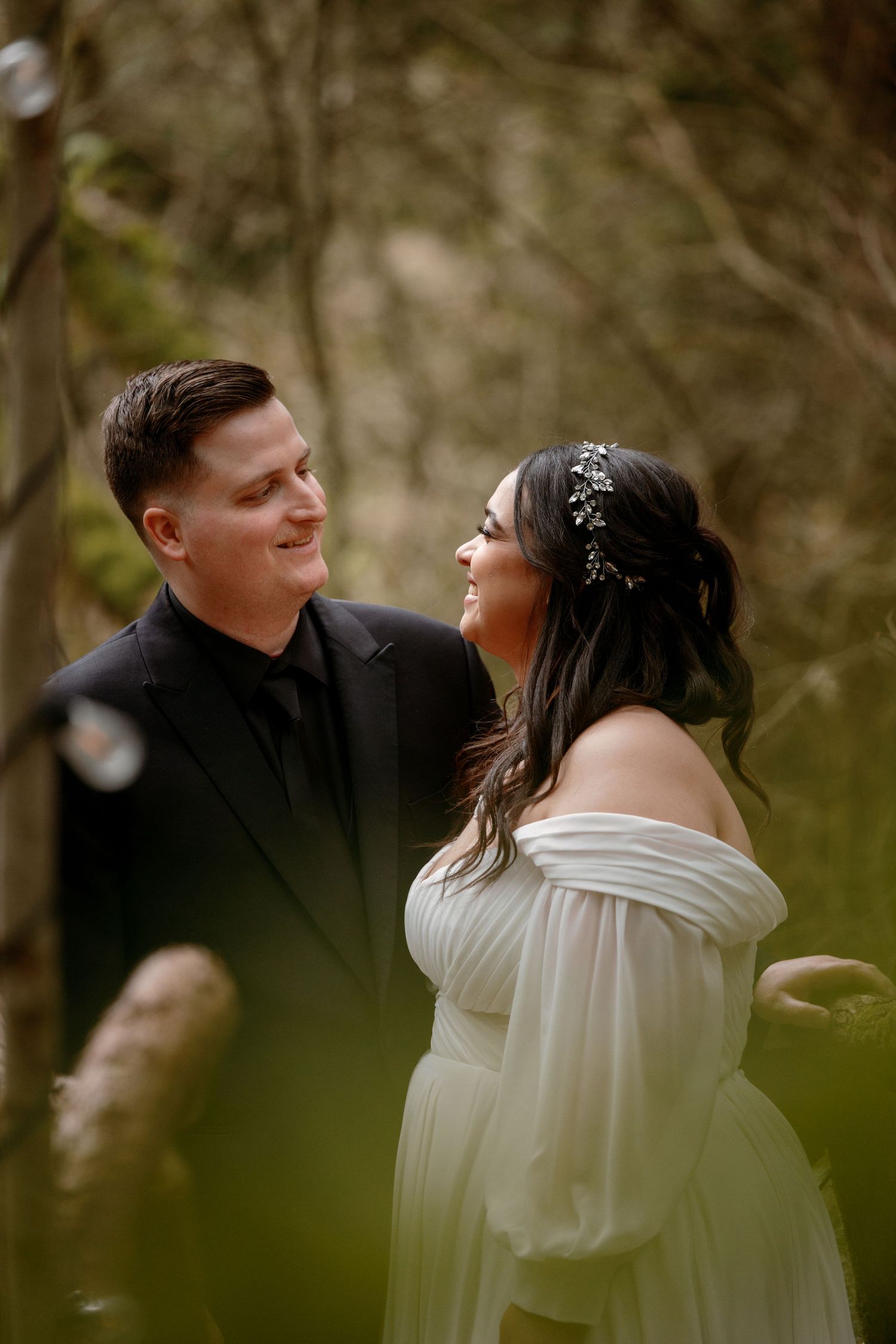 Smiling bride and groom gazing at each other lovingly among soft bokeh greenery at forest wedding.