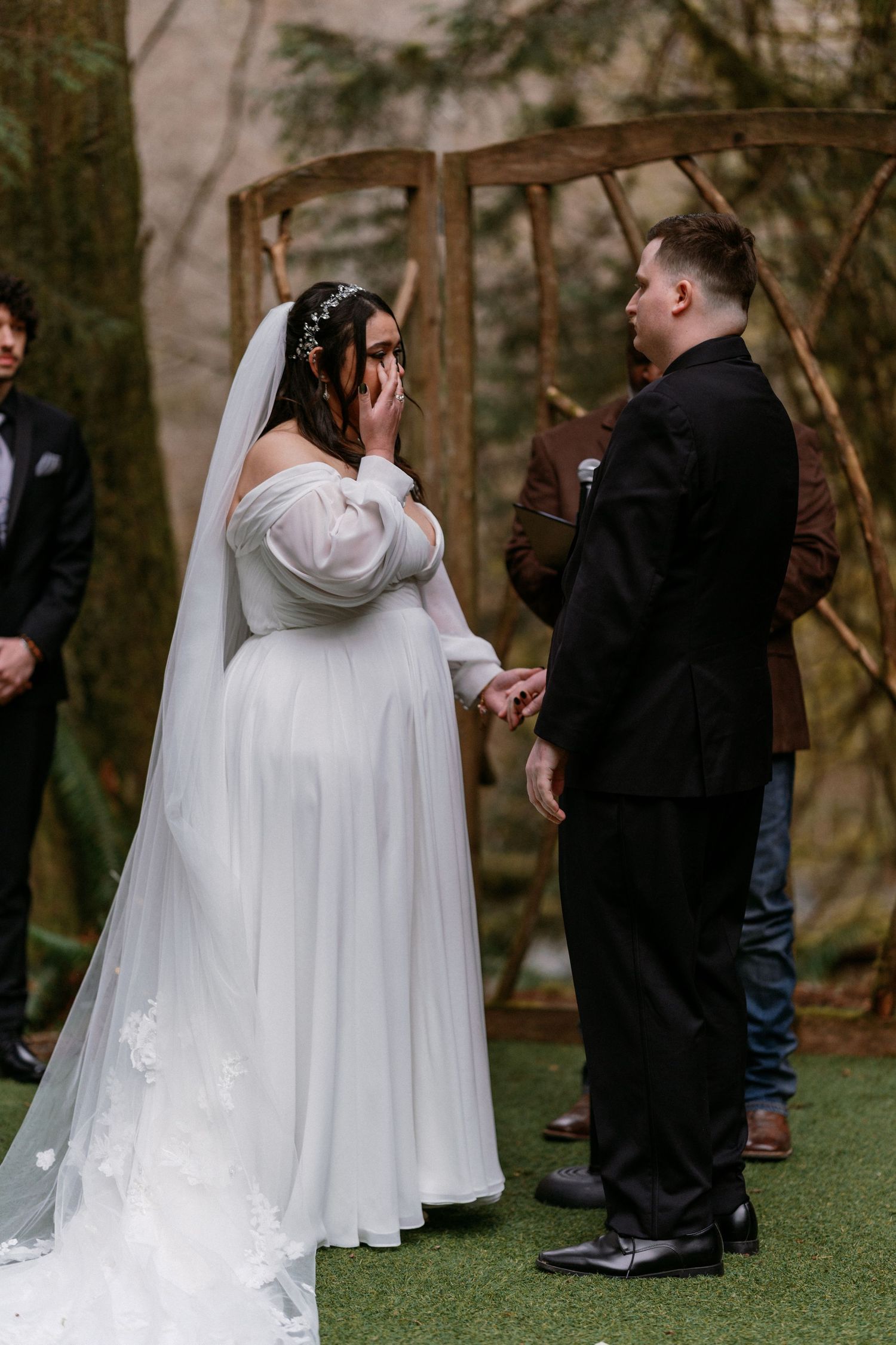 Emotional bride in white off-shoulder gown wiping tears during outdoor forest wedding ceremony.