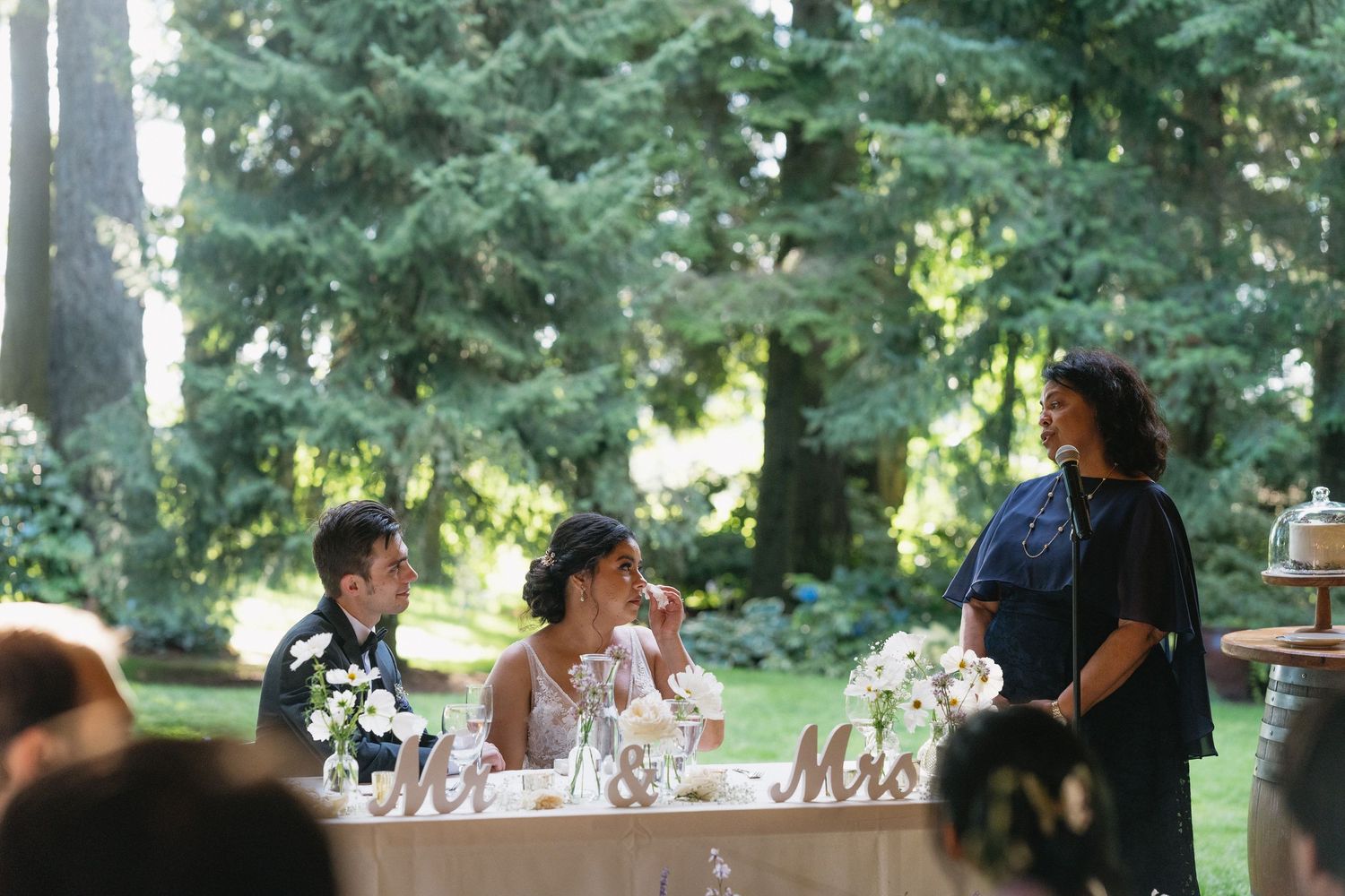 Wedding guest giving speech to smiling bride and groom at outdoor Mr. and Mrs. sweetheart table.