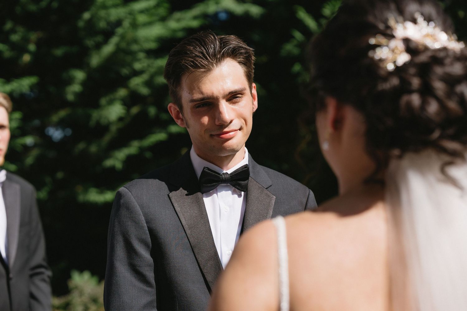 Groom in gray suit and bow tie gazes lovingly at bride during outdoor garden wedding ceremony.