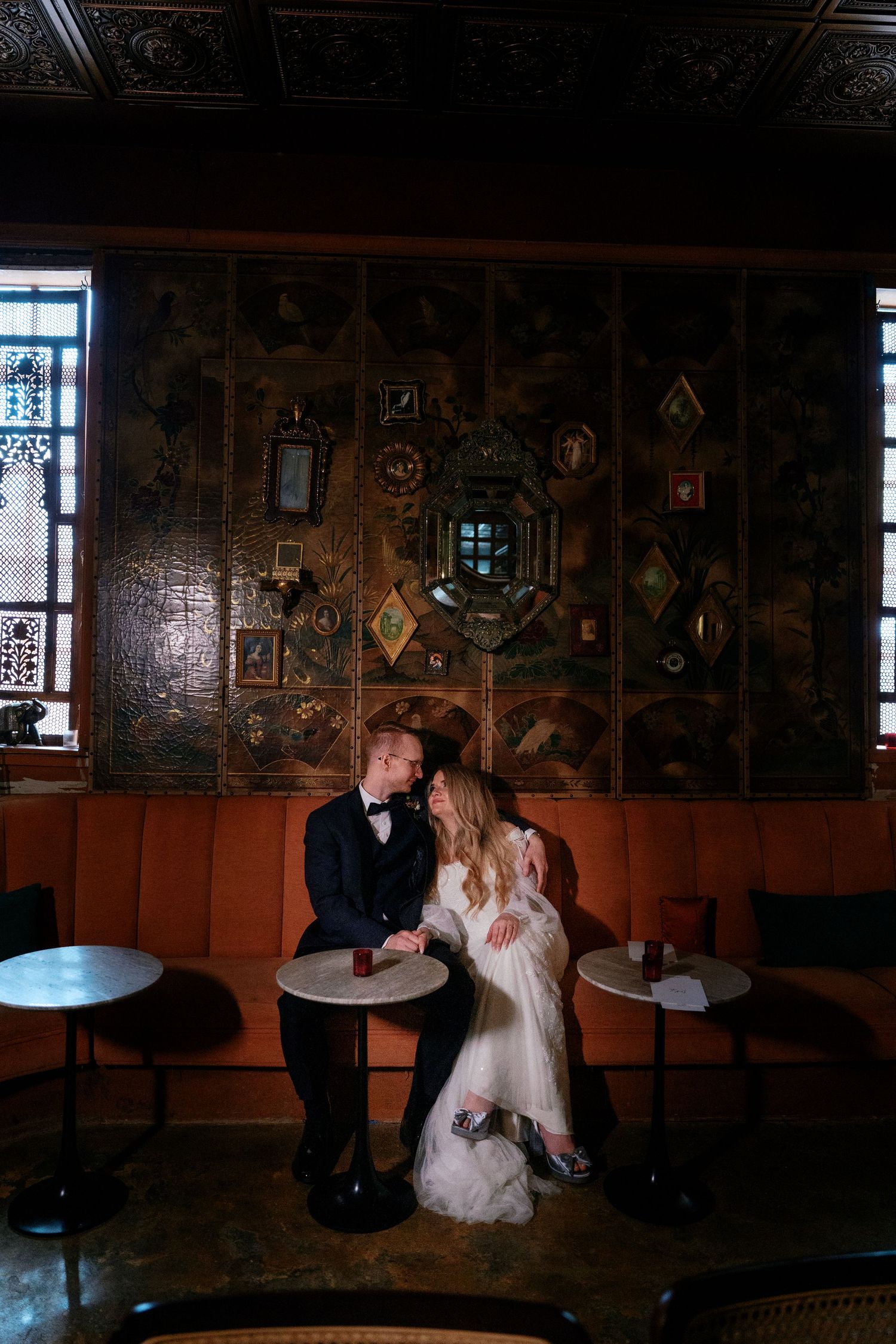 Bride and groom share intimate moment leaning close at small table in elegant dark wood-paneled bar with stained glass windows.